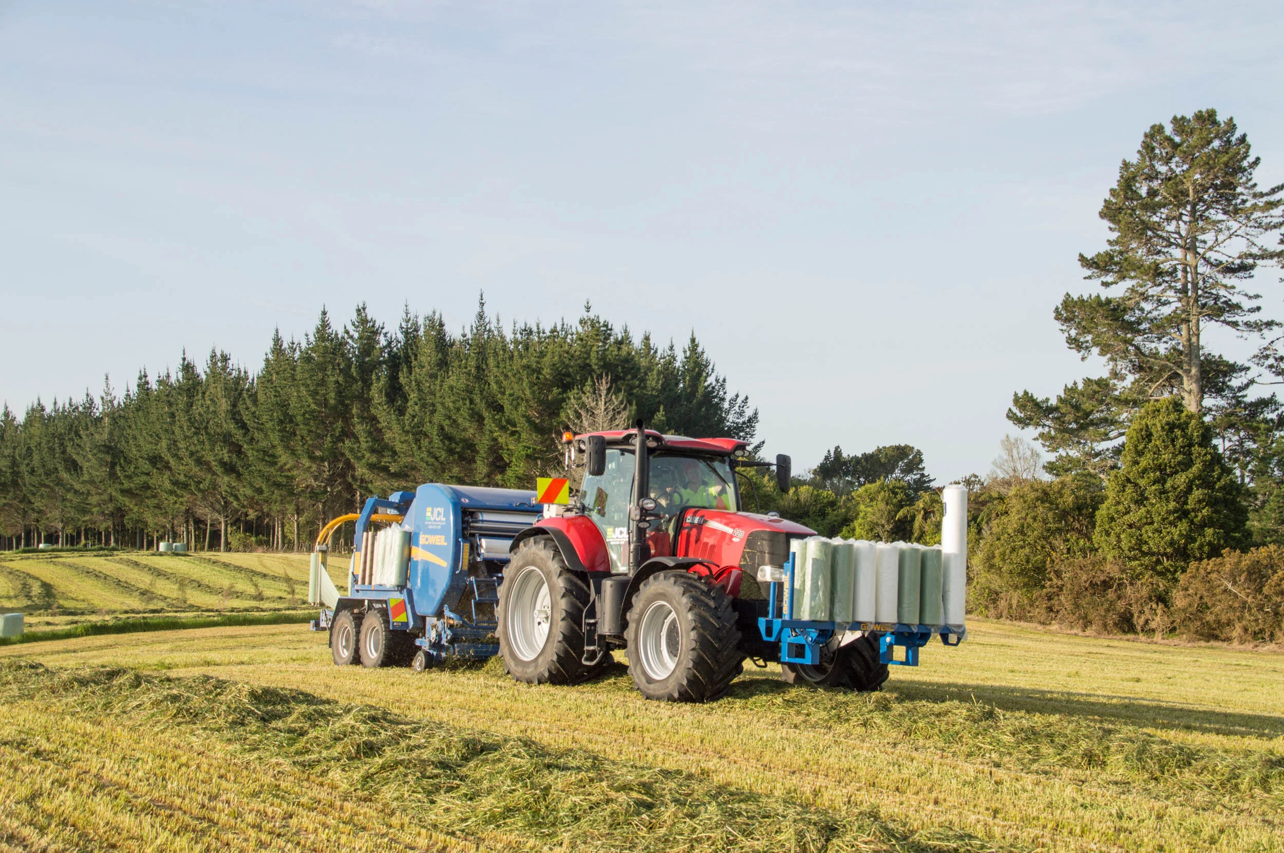 A red tractor using machinery on a field with trees in the background.