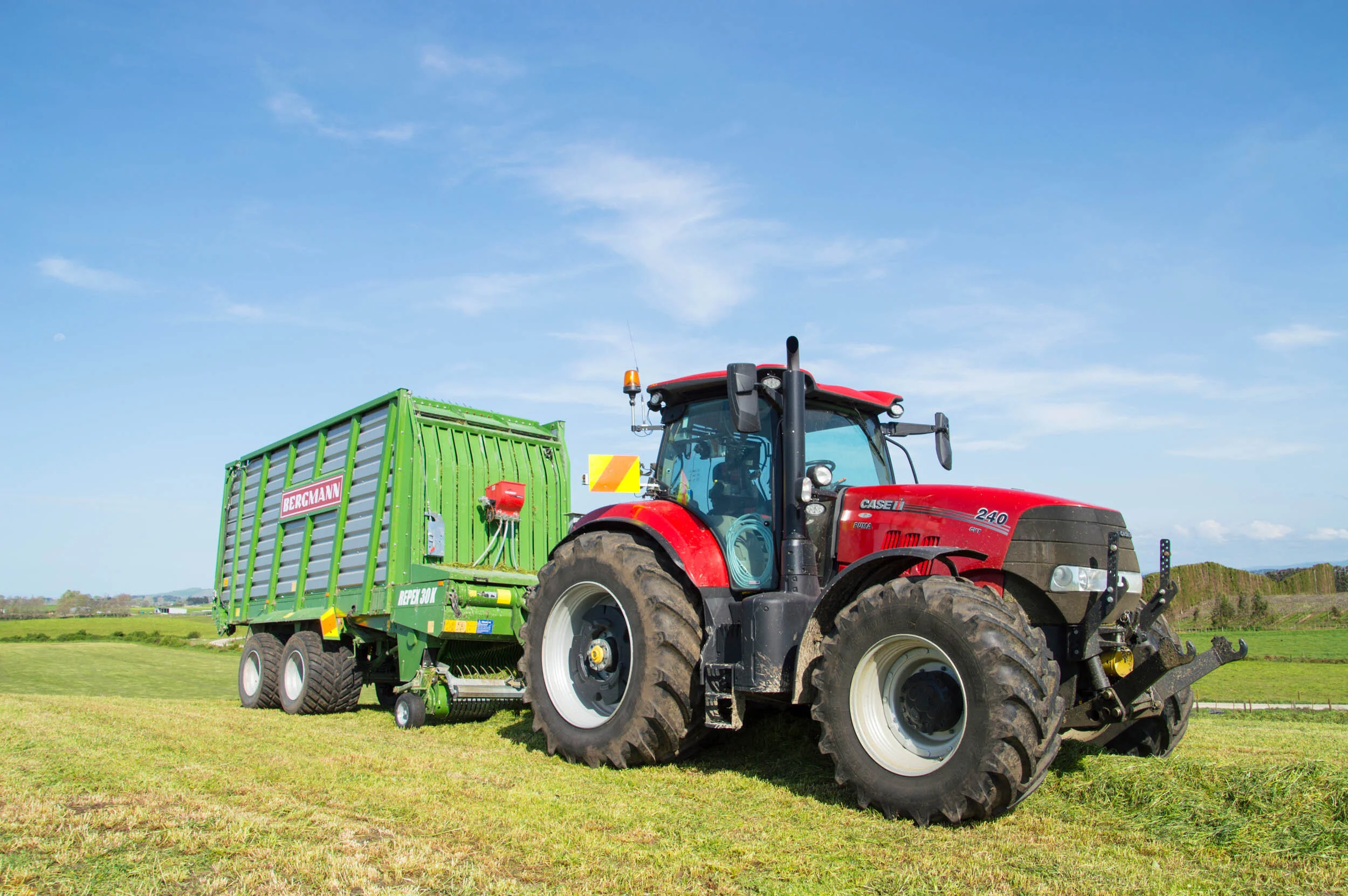 Red tractor pulling a green bergmann manure spreader on a grassy field under a blue sky.