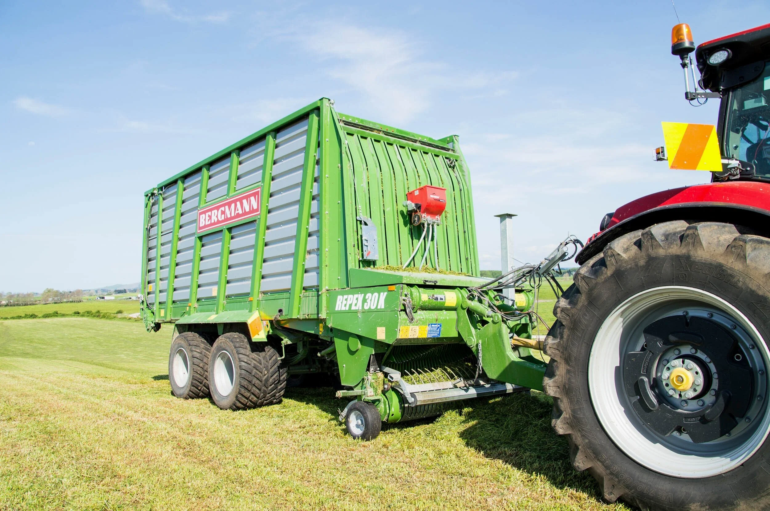 A green Bergmann Repex 30 M hay baler attached to a red tractor on a grassy field under a blue sky with some clouds.