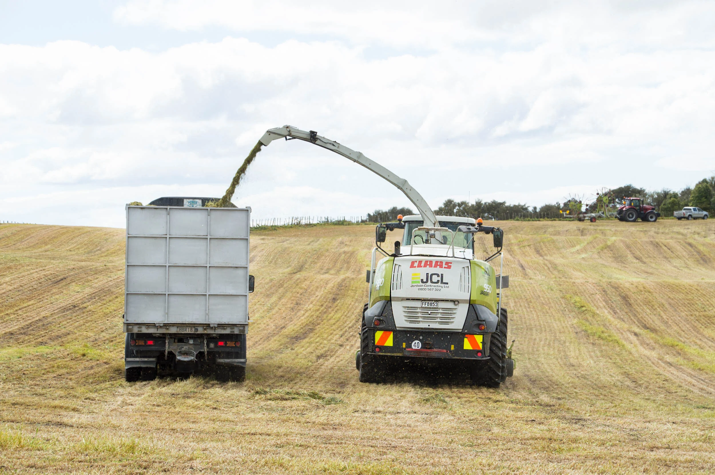 A piece of farm machinery collects grass from a field, with two tractors and a vehicle in the background under a cloudy sky.
