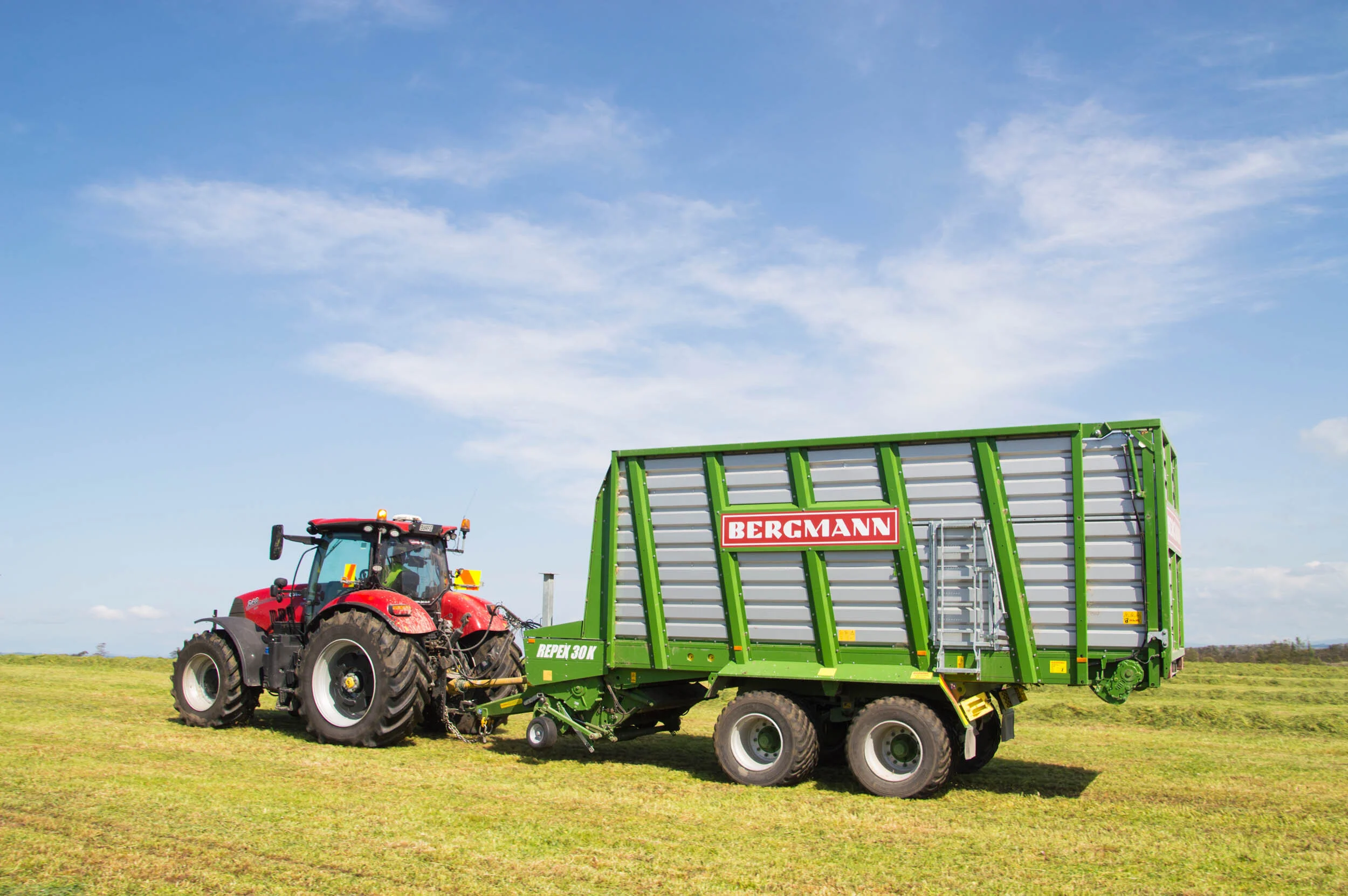 Red tractor attached to a green Bergmann forage wagon on a green field under a partly cloudy sky.
