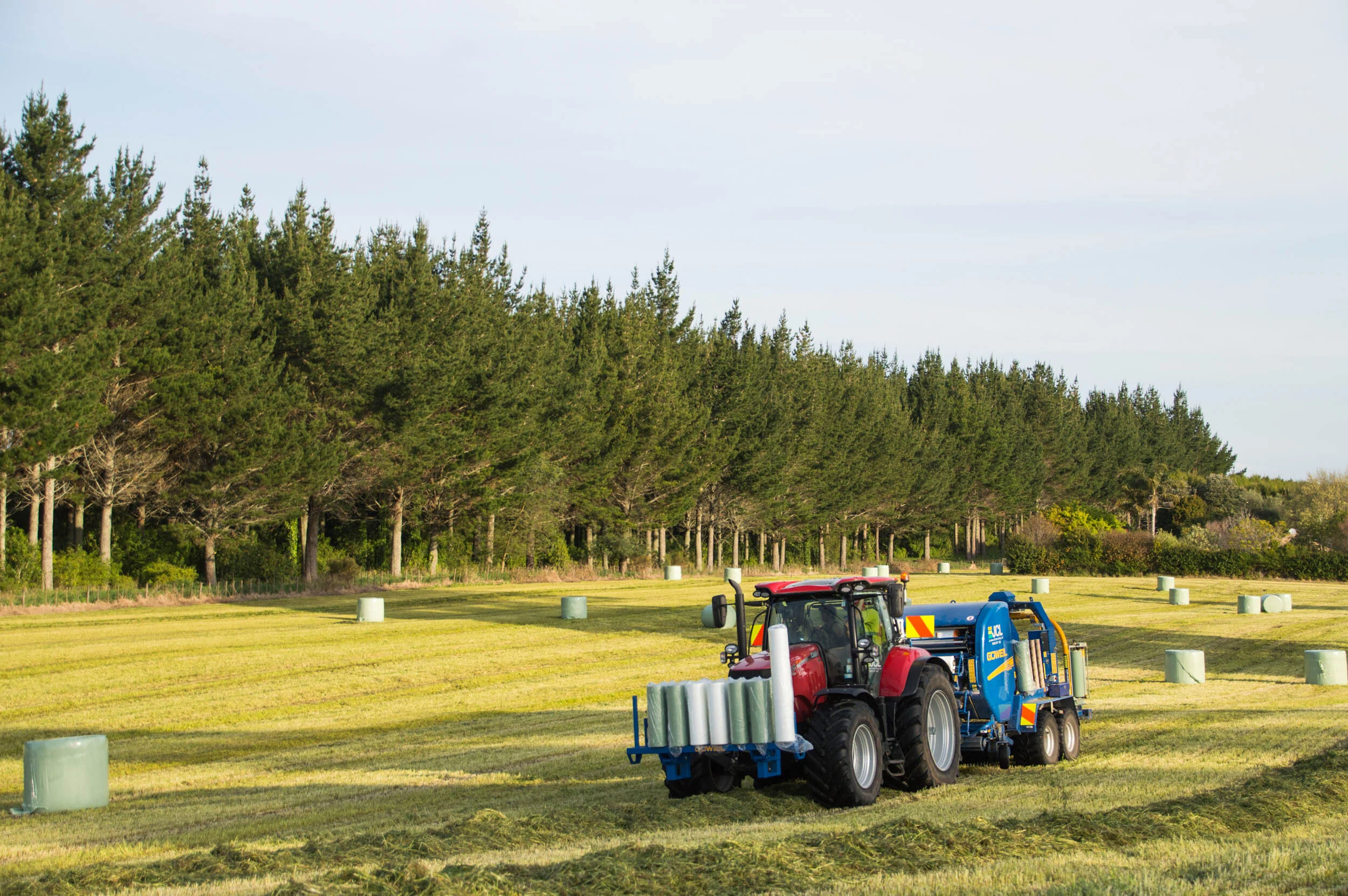 Red tractor pulling a blue applicator machine on a grassy field with hay bales and trees in the background.