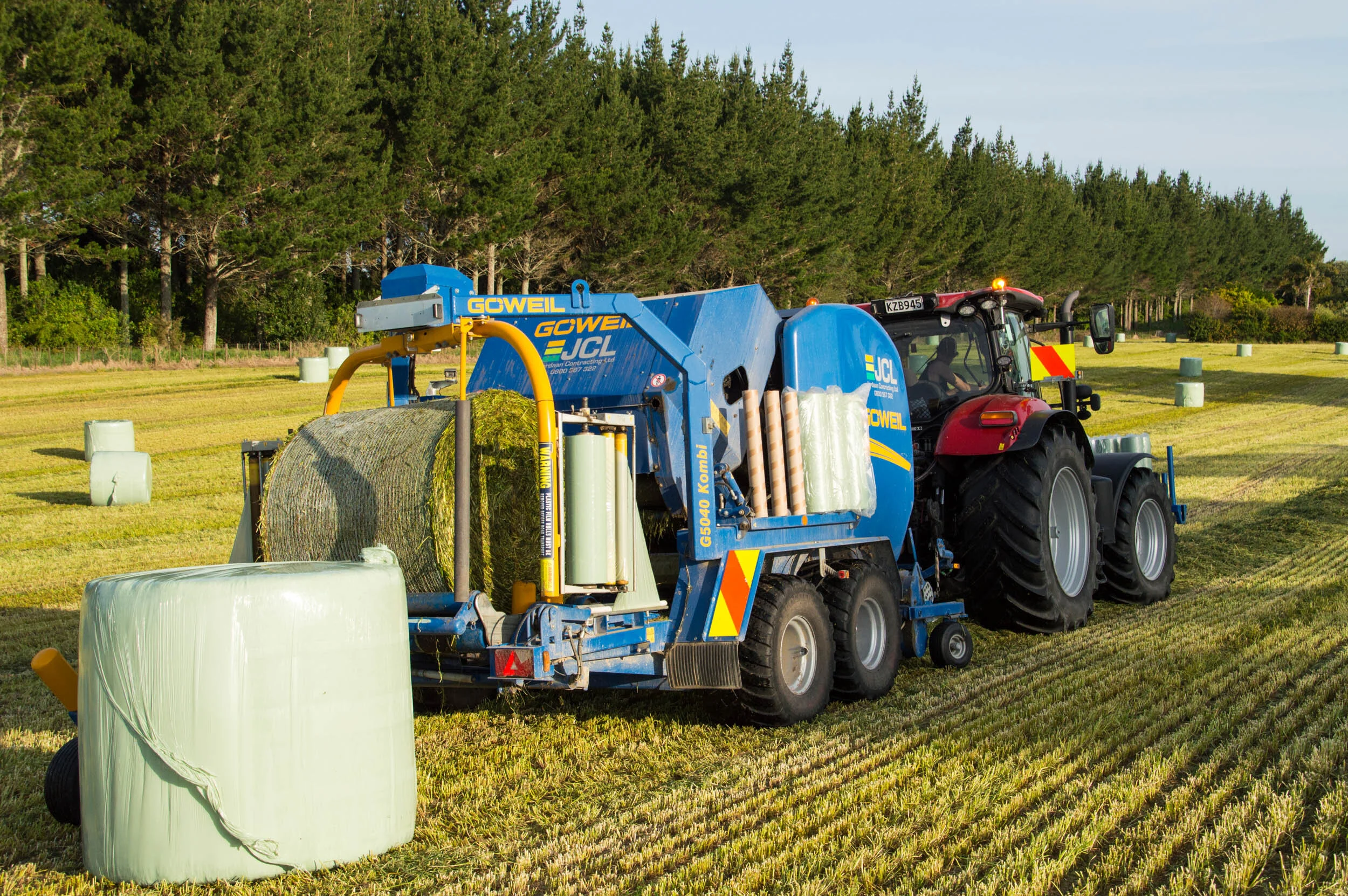 A tractor with a large baler attachment working in a field, creating round hay bales during daytime.