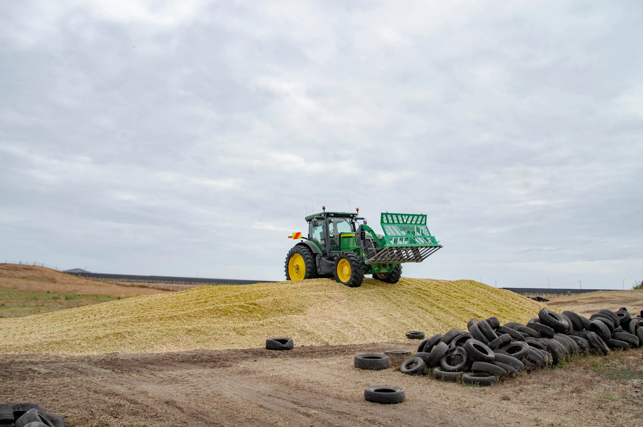 A green tractor with yellow wheels on a dirt hill, carrying a green crate. Piles of black tires are scattered in the foreground, with a cloudy sky overhead.