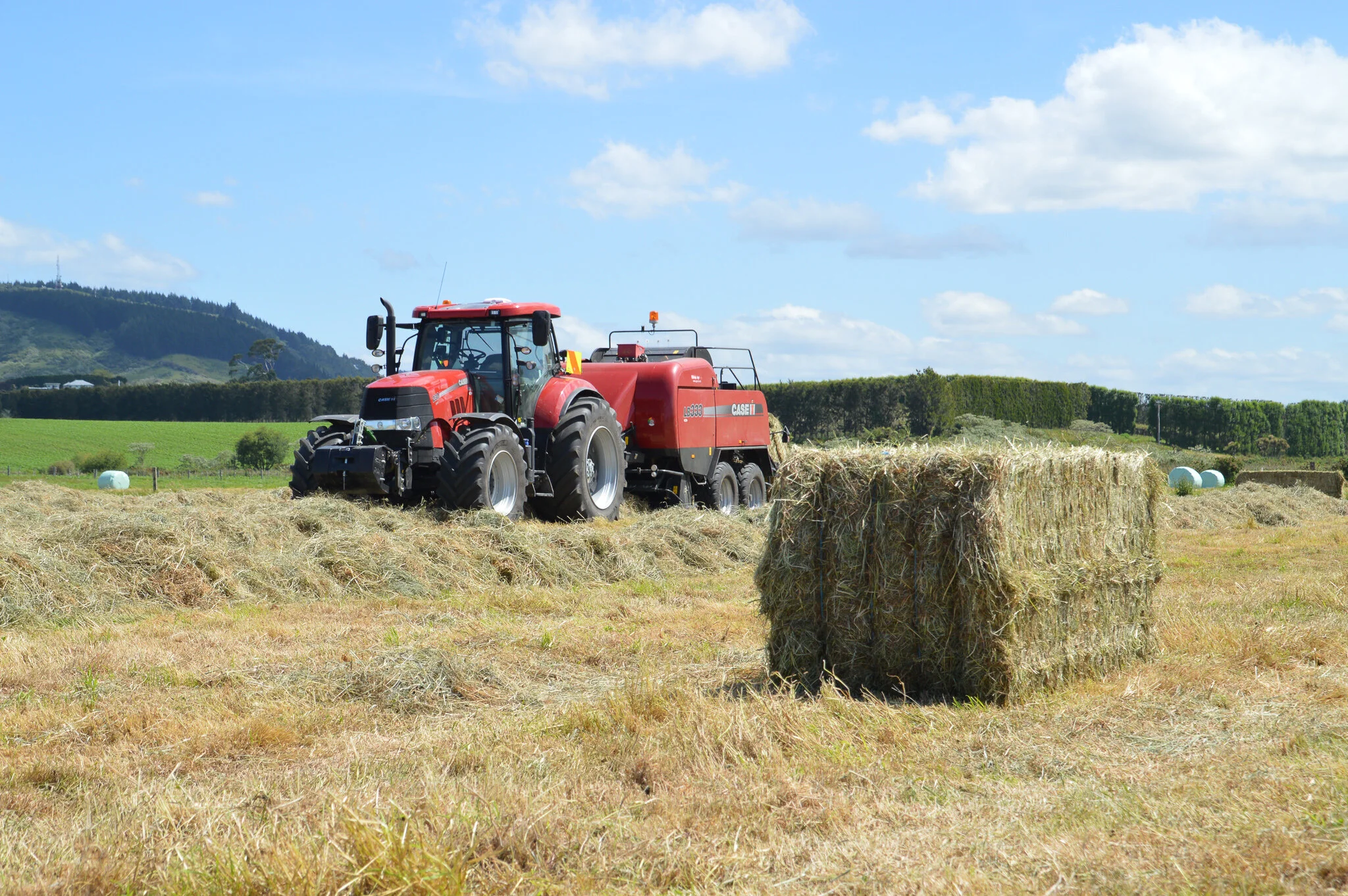 Red tractor moving hay on a field with a large hay bale in the foreground, under a partly cloudy sky.