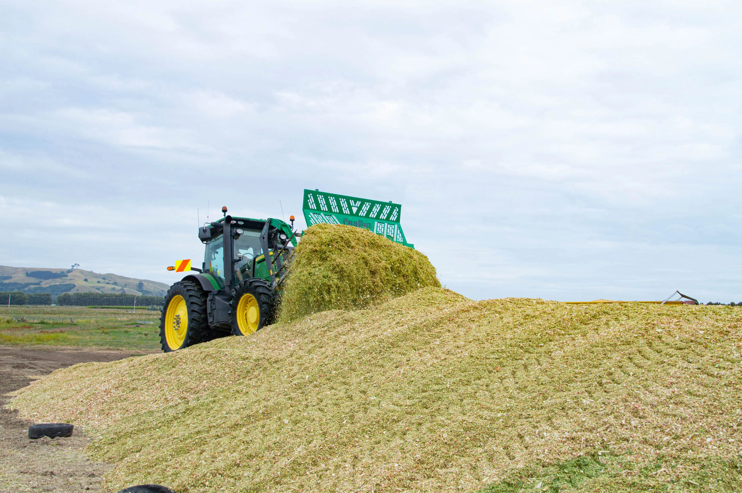 A green tractor spreading harvested grain onto a pile on a farm field under a cloudy sky.