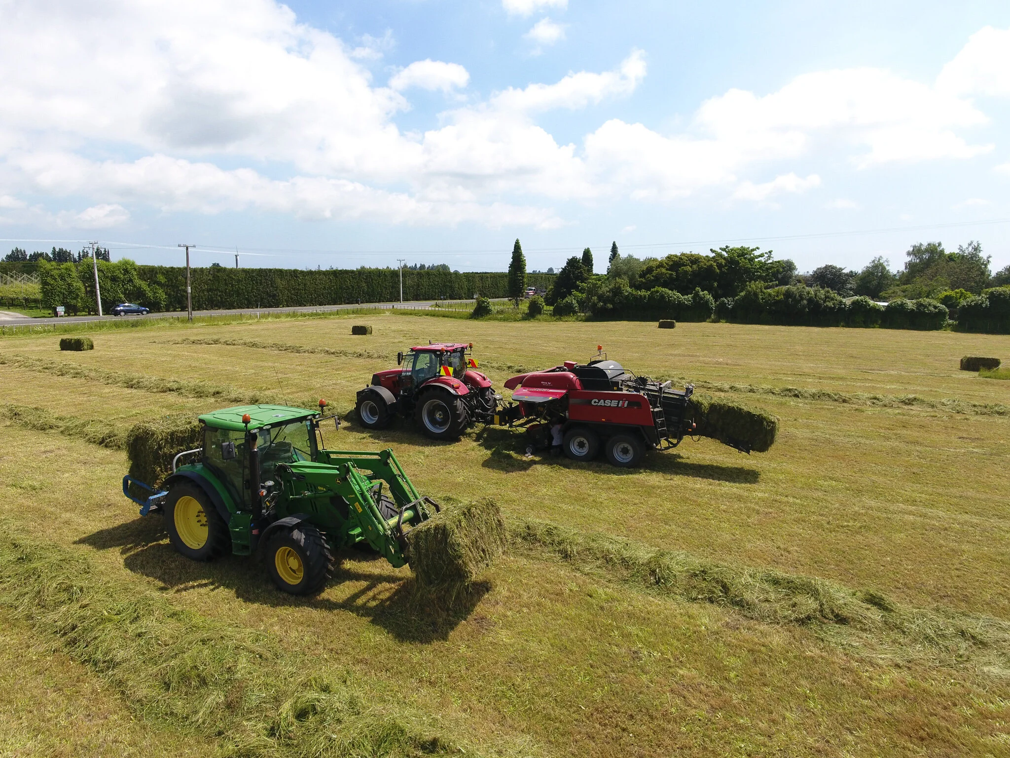 Three tractors harvesting hay in a field with scattered hay bales, blue sky with clouds, and trees in the background.