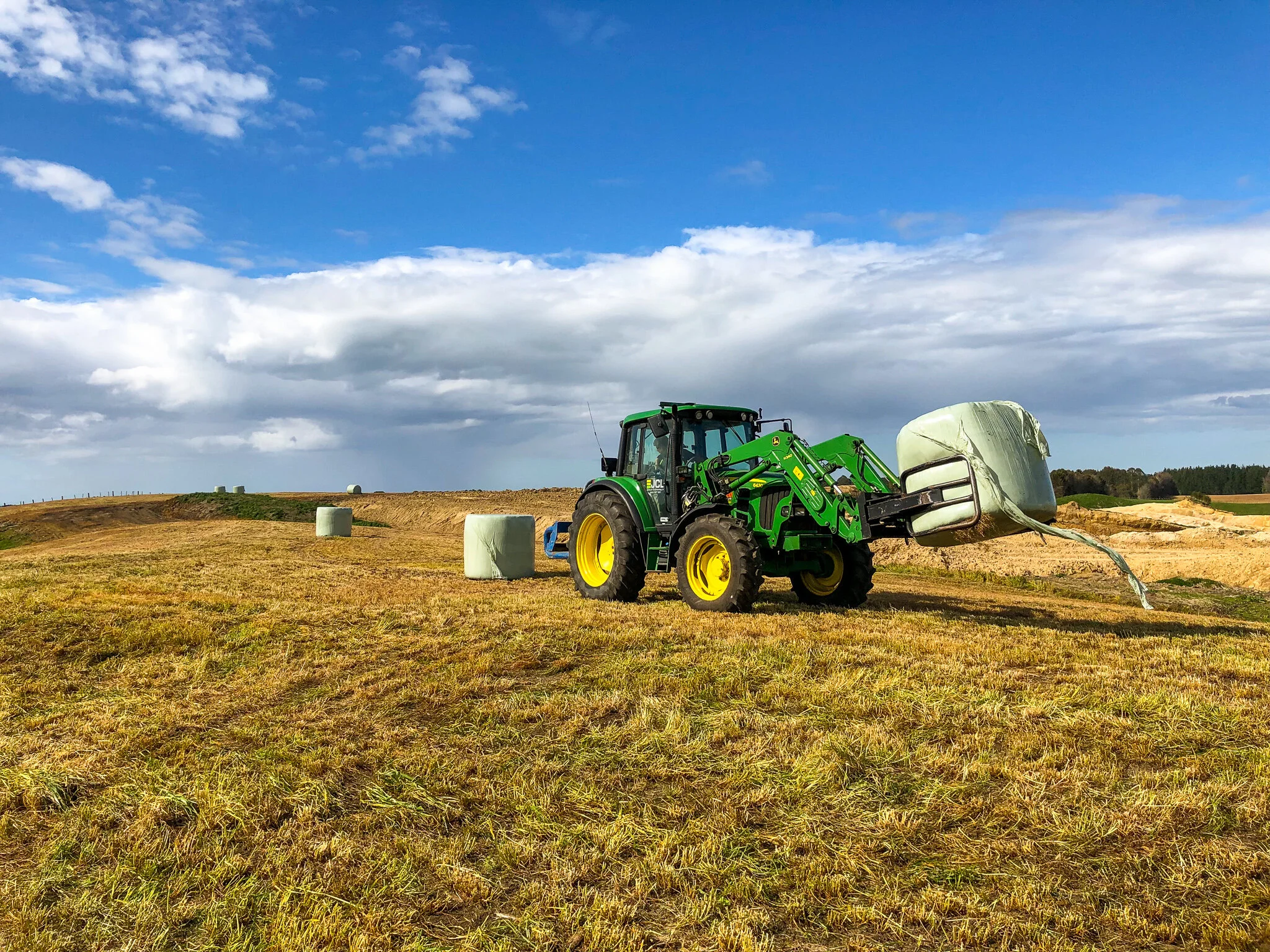 A green tractor with yellow tires lifting a large hay bale in a farm field under a blue sky with scattered clouds.