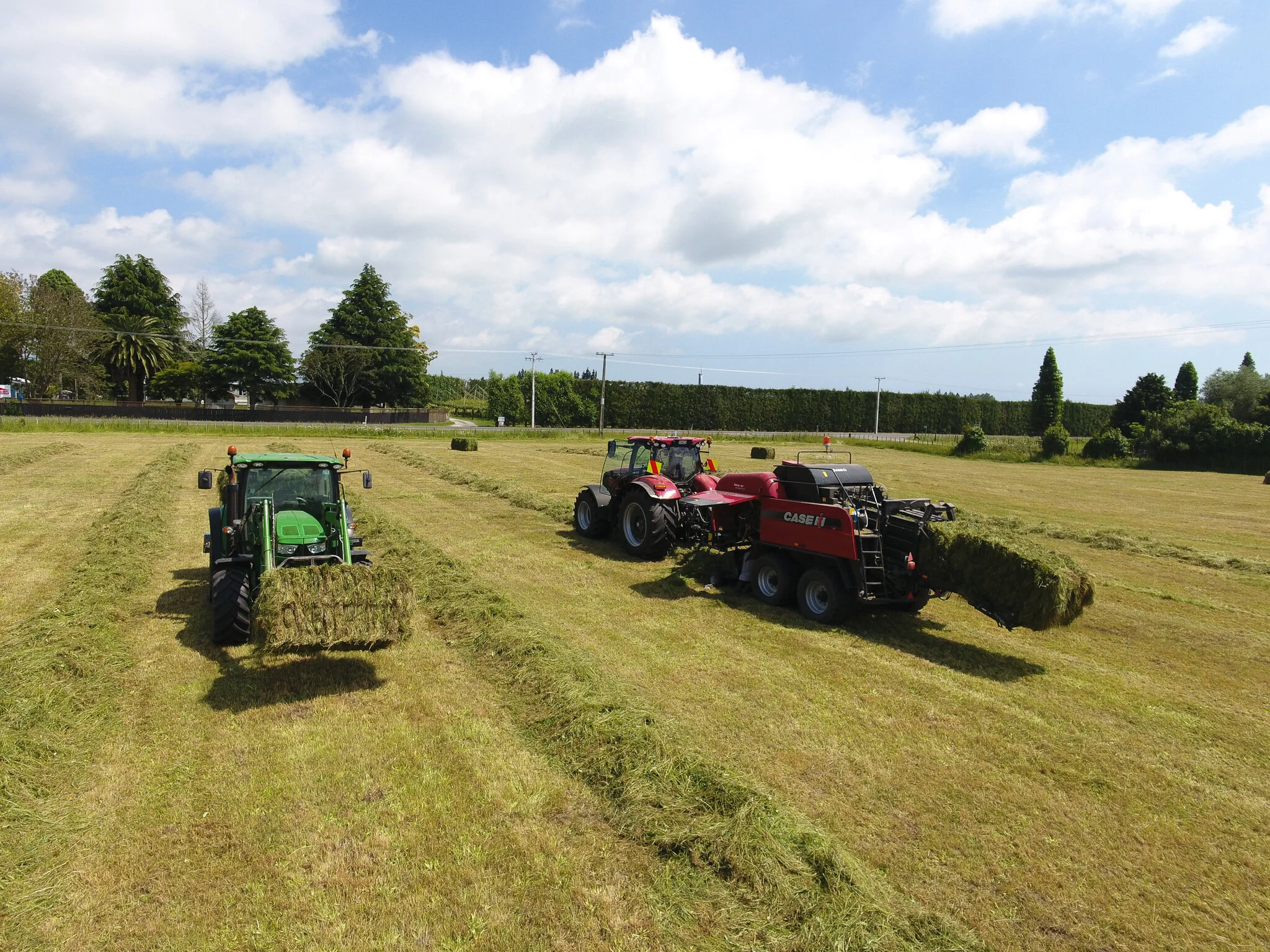 Two tractors, one green and one red, are harvesting hay in a large field under a partly cloudy sky.