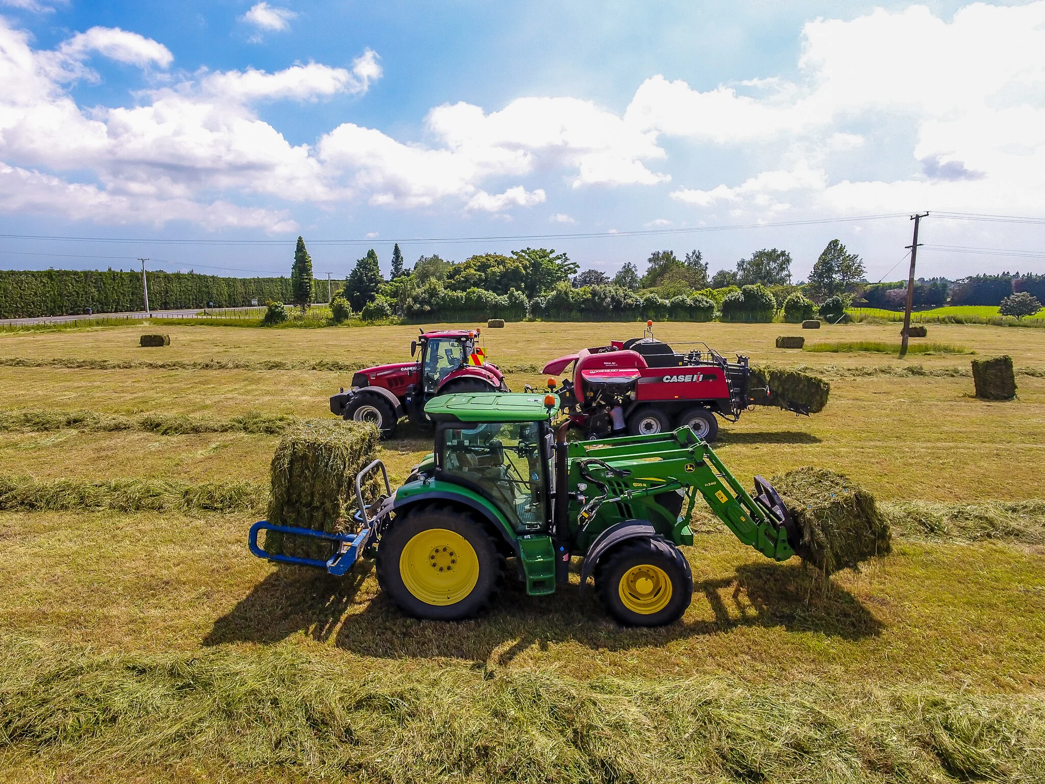Two tractors, one green and one red, are in a field with hay bales, harvesting or moving hay on a sunny day with a partly cloudy sky.