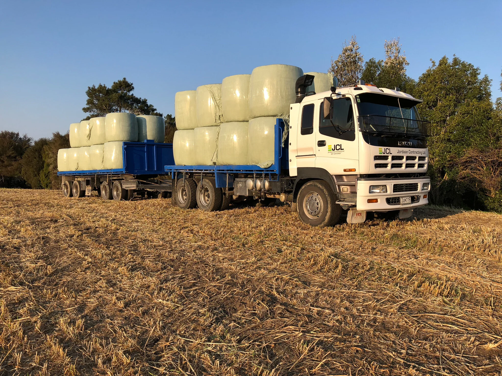 A large flatbed truck loaded with hay bales on a farmland with trees in the background.
