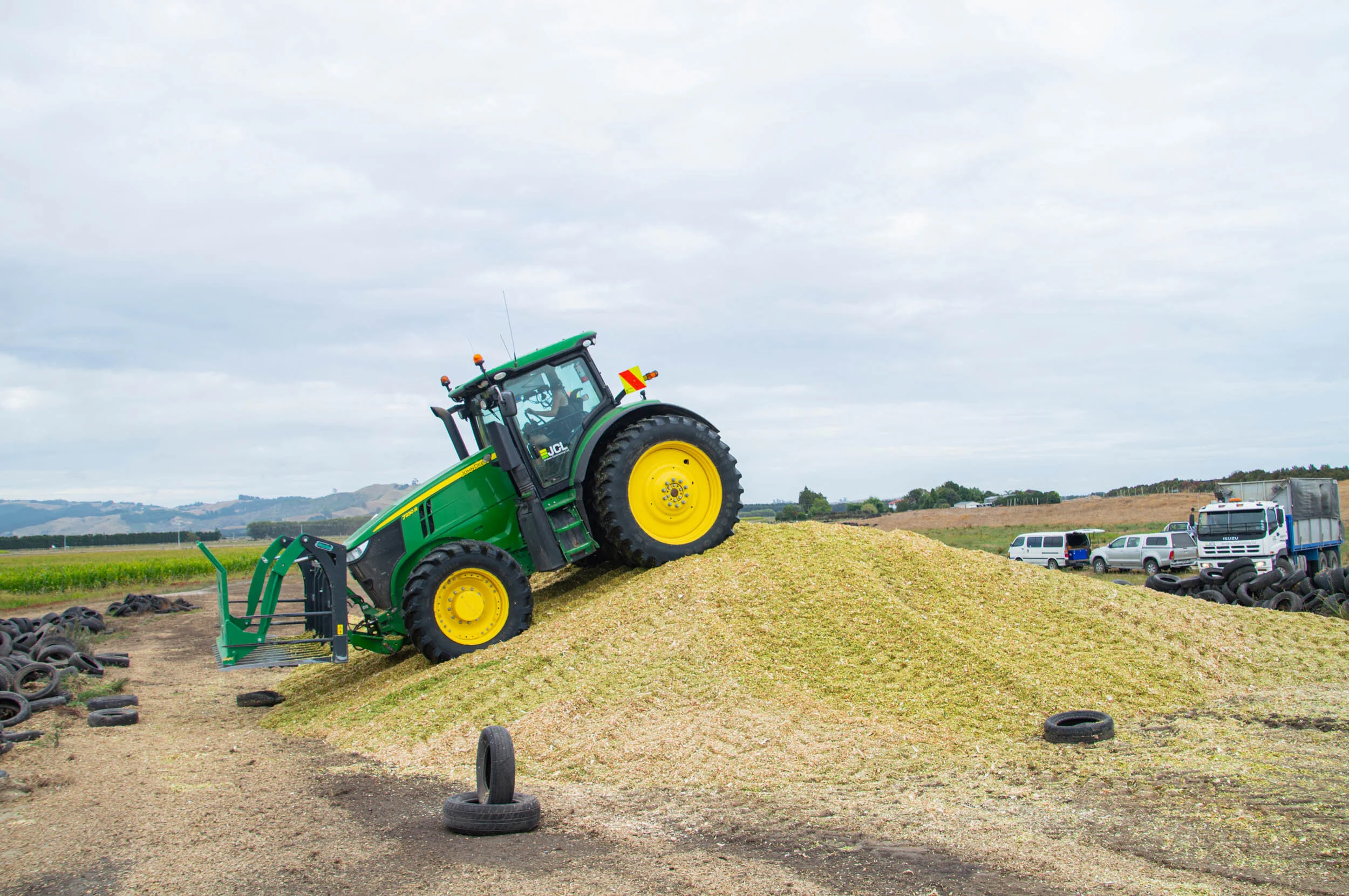 A green tractor on a mound of harvested crop residue at a farm field, with tires and trucks in the background.