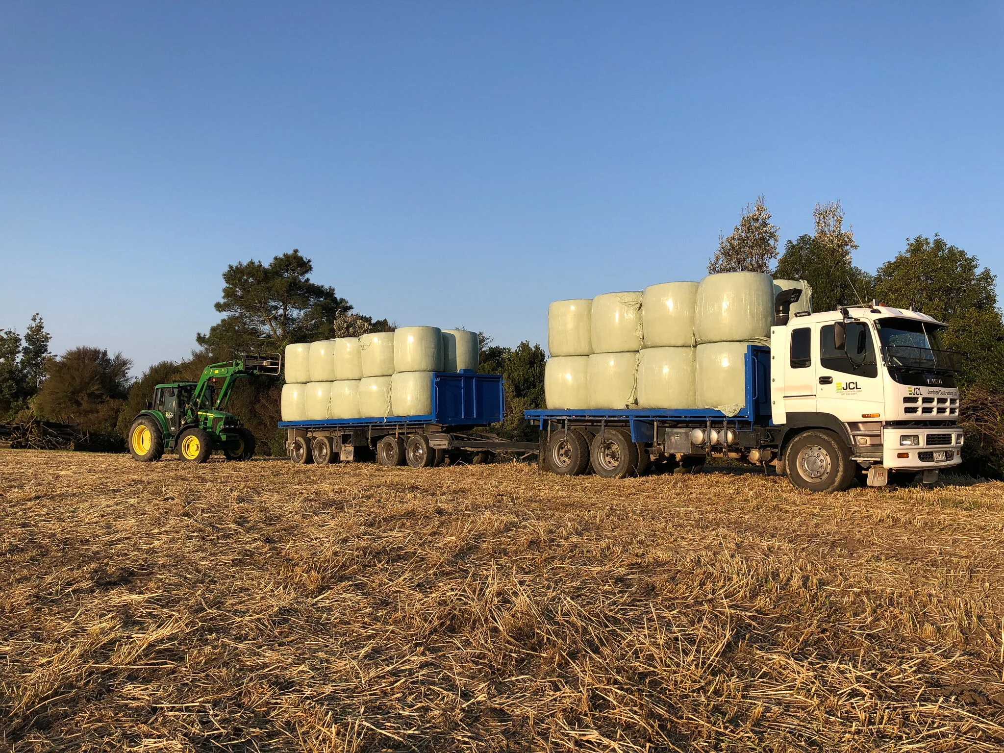 A white truck with blue sides carrying large wrapped bales of hay, being unloaded by a green tractor with a front loader on a farm field under a clear blue sky.