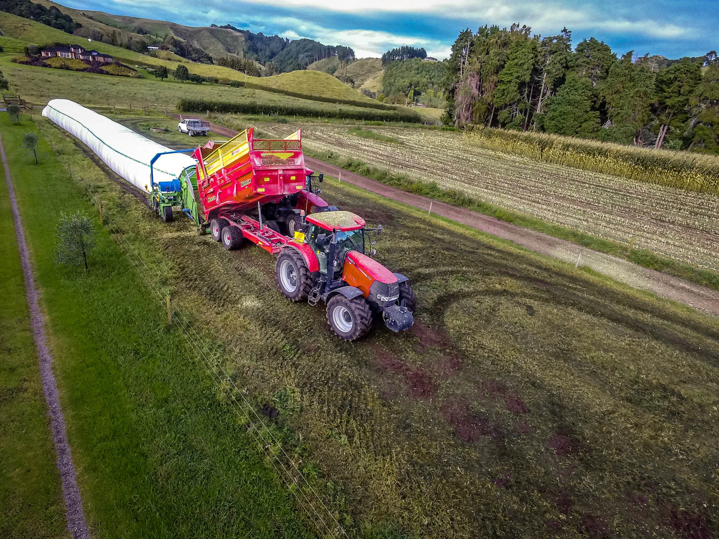 A tractor with a pink and orange color scheme is hauling a pink manure spreader in a rural farmland. The spreader is releasing a greenish substance onto the field as part of land management. The landscape features rolling green hills, trees, and cult