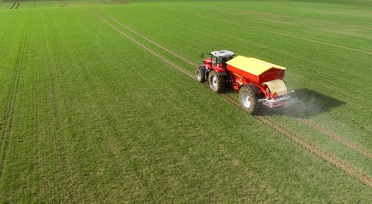A red tractor with a yellow attachment spreading fertilizer or seeds on a green field.