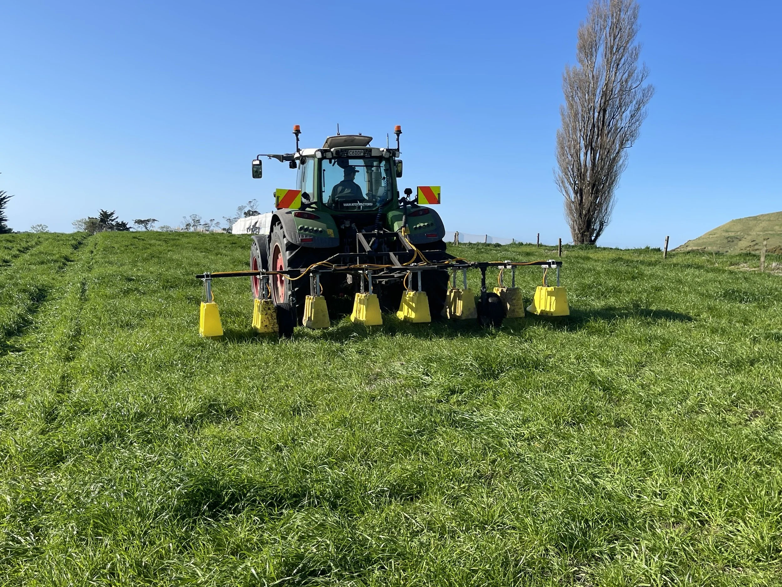 A green tractor with yellow spray nozzles attached is spraying a crop field under a clear blue sky, with trees and hills in the background.