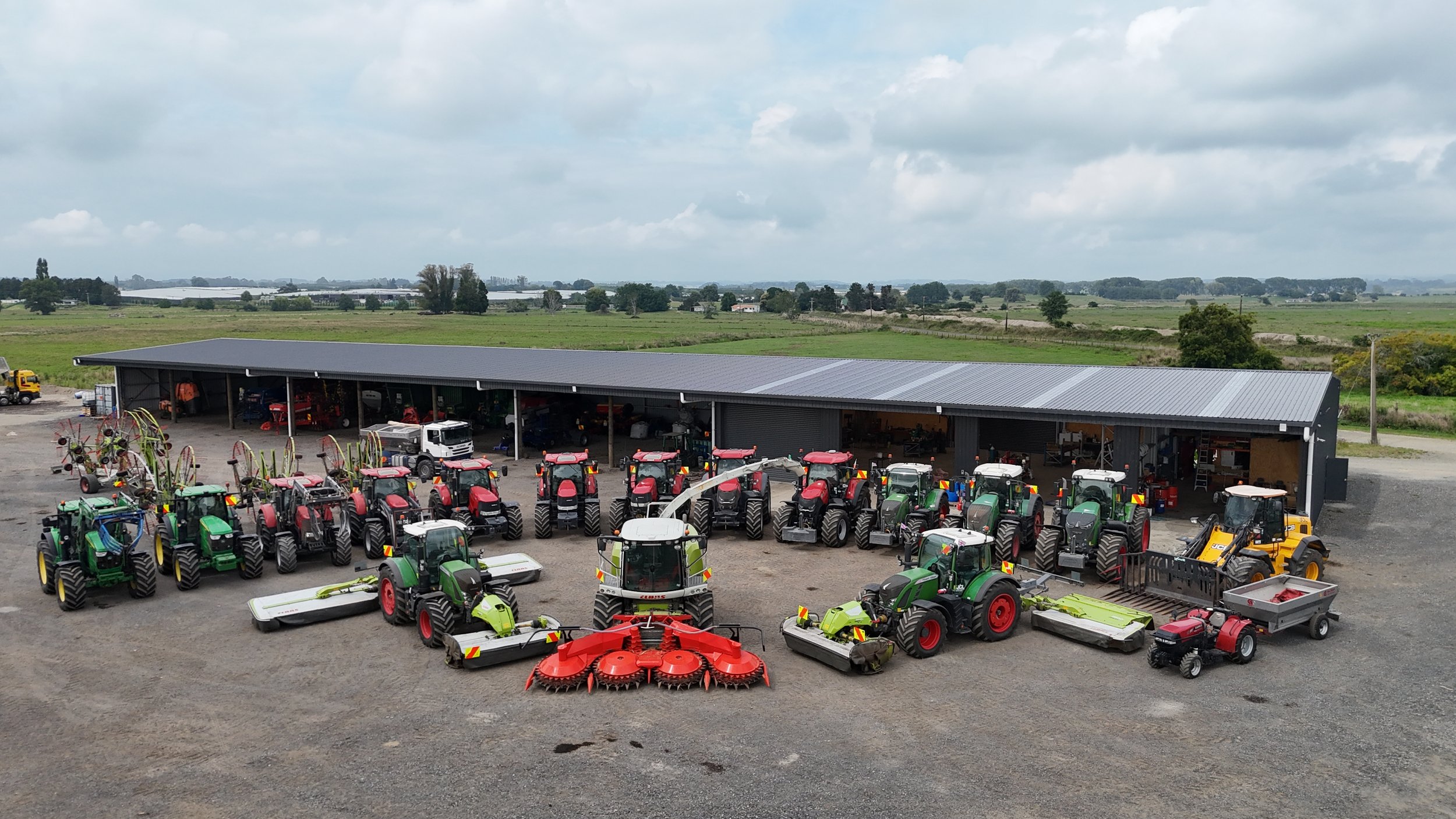 An outdoor display of various tractors and agricultural equipment is arranged in front of a large metal shed on a farm. The tractors are mostly red and green, with some yellow equipment and a few trucks visible. The background shows open fields and a partly cloudy sky.