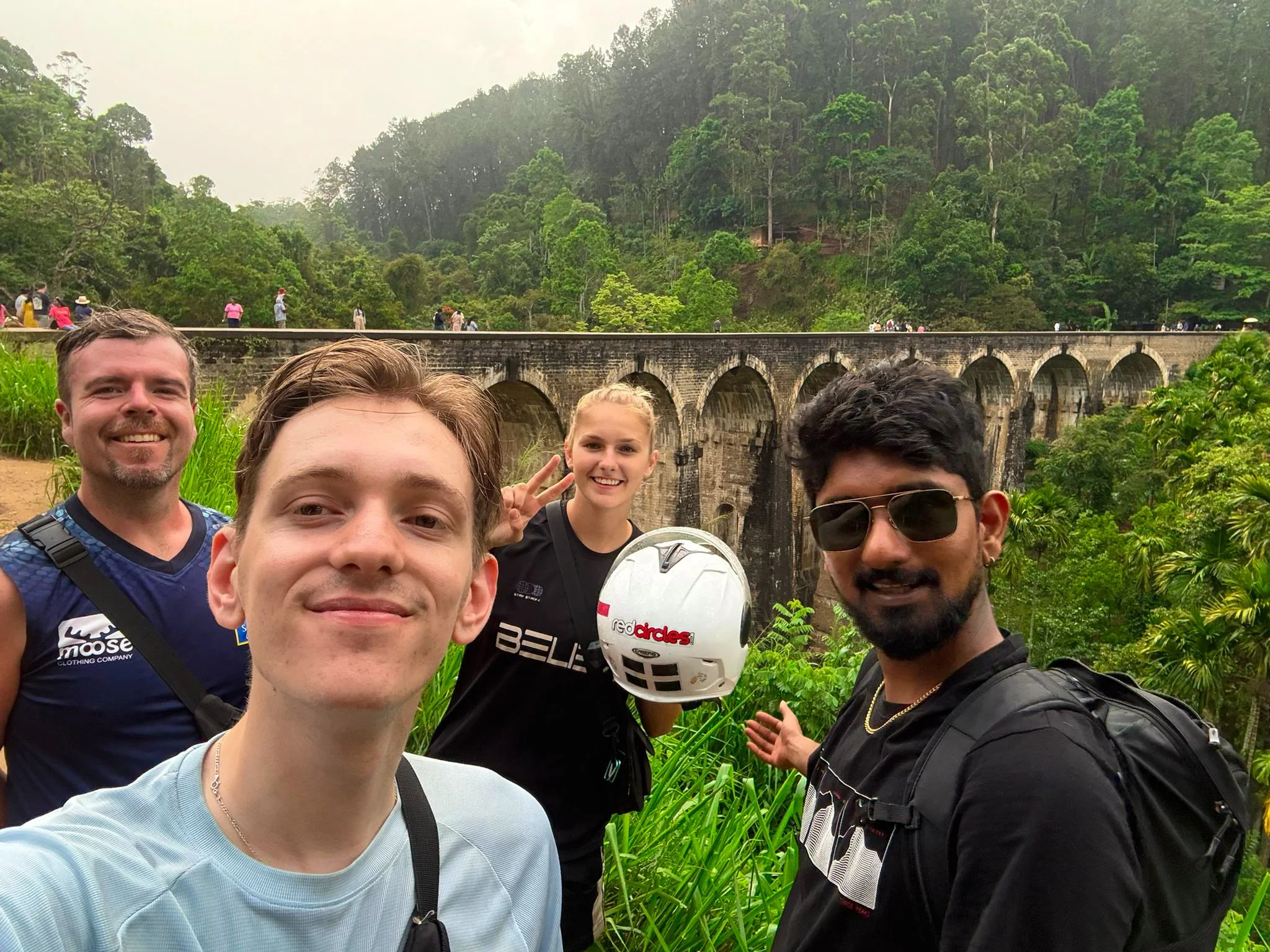 Group of six people taking a selfie outdoors in a green, forested area with a stone bridge and lush trees in the background.