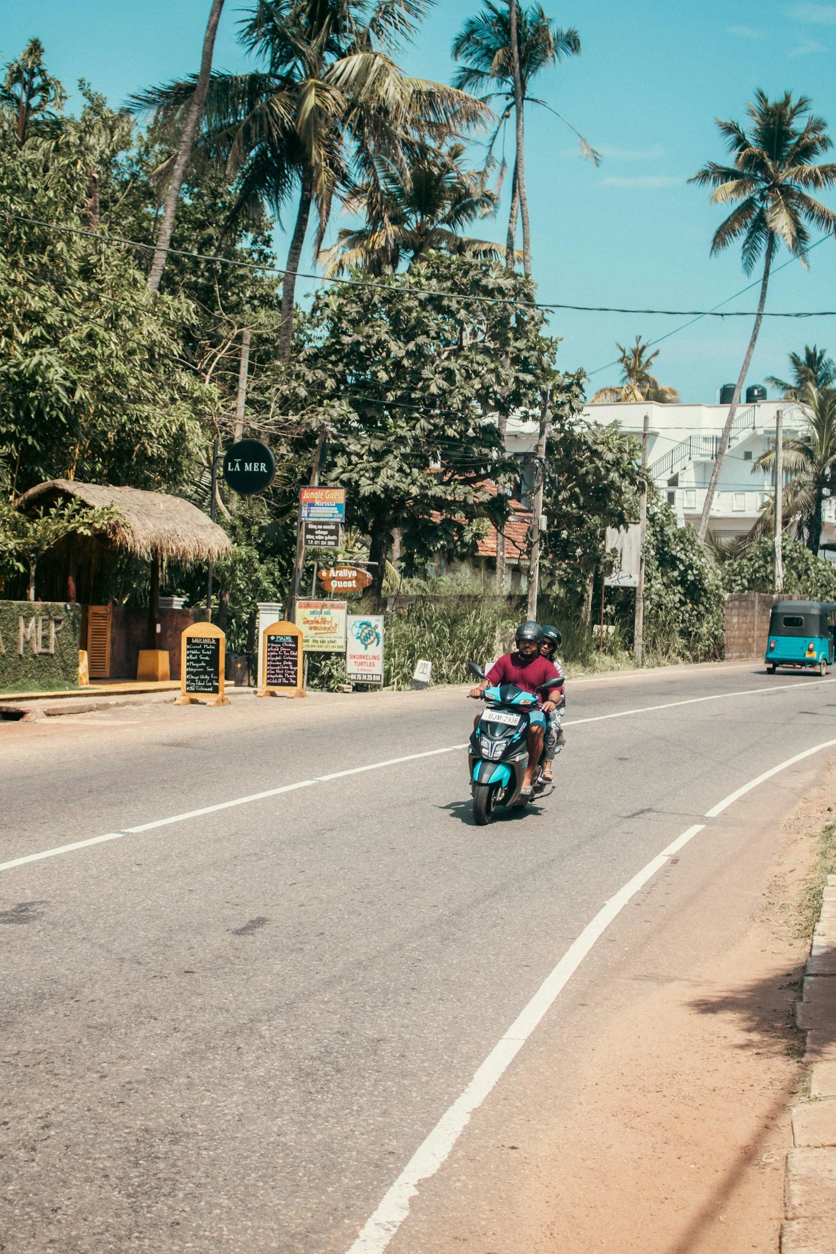 A person riding a blue scooter on a curved road with sandy shoulder. There are lush green trees, including palm trees, and signage for local businesses nearby. A person wearing a helmet and sunglasses is driving the scooter during the daytime.