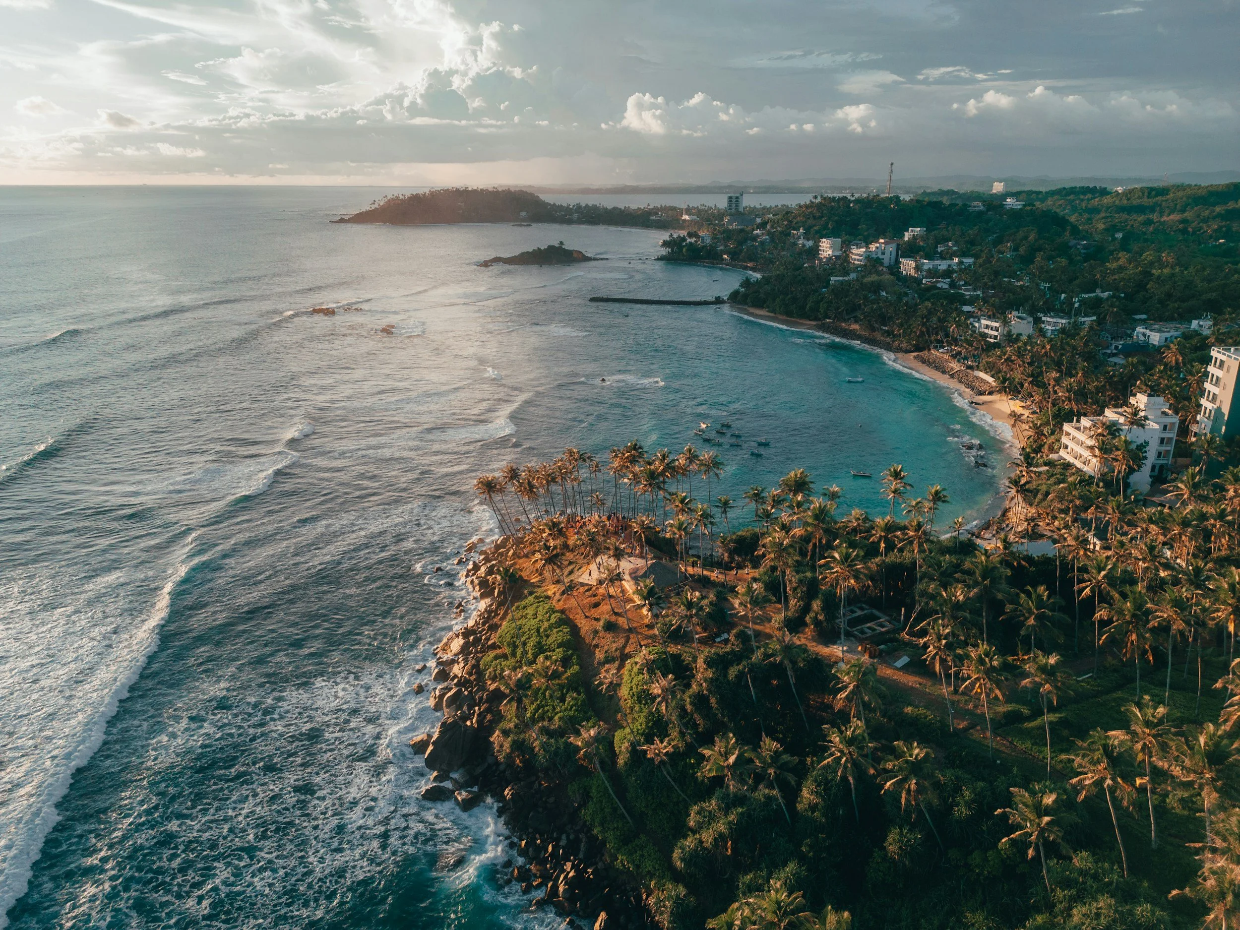 Aerial view of a tropical coastal area with a beach, palm trees, and buildings on a hillside. The ocean waves are crashing onto the shoreline under a cloudy sky.