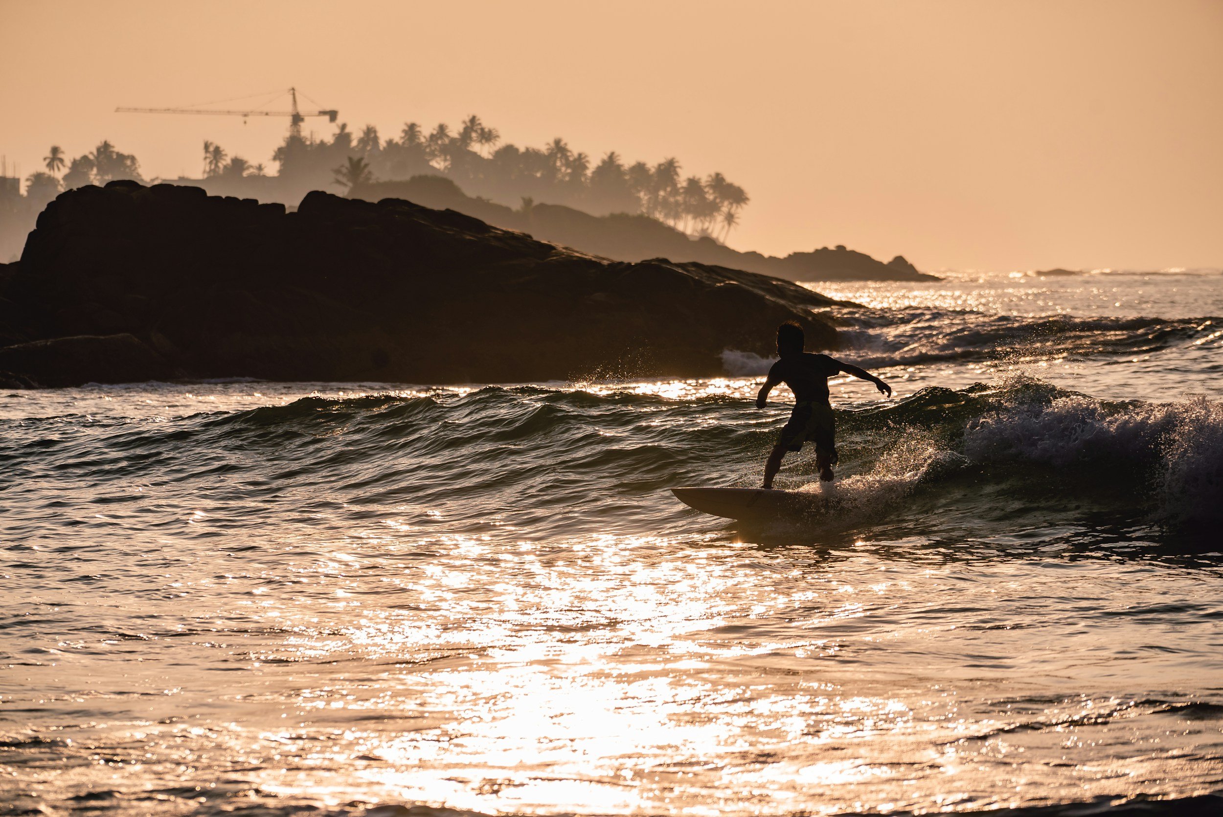 A silhouette of a person surfing in the ocean during sunset with rocky shoreline and palm trees in the background.