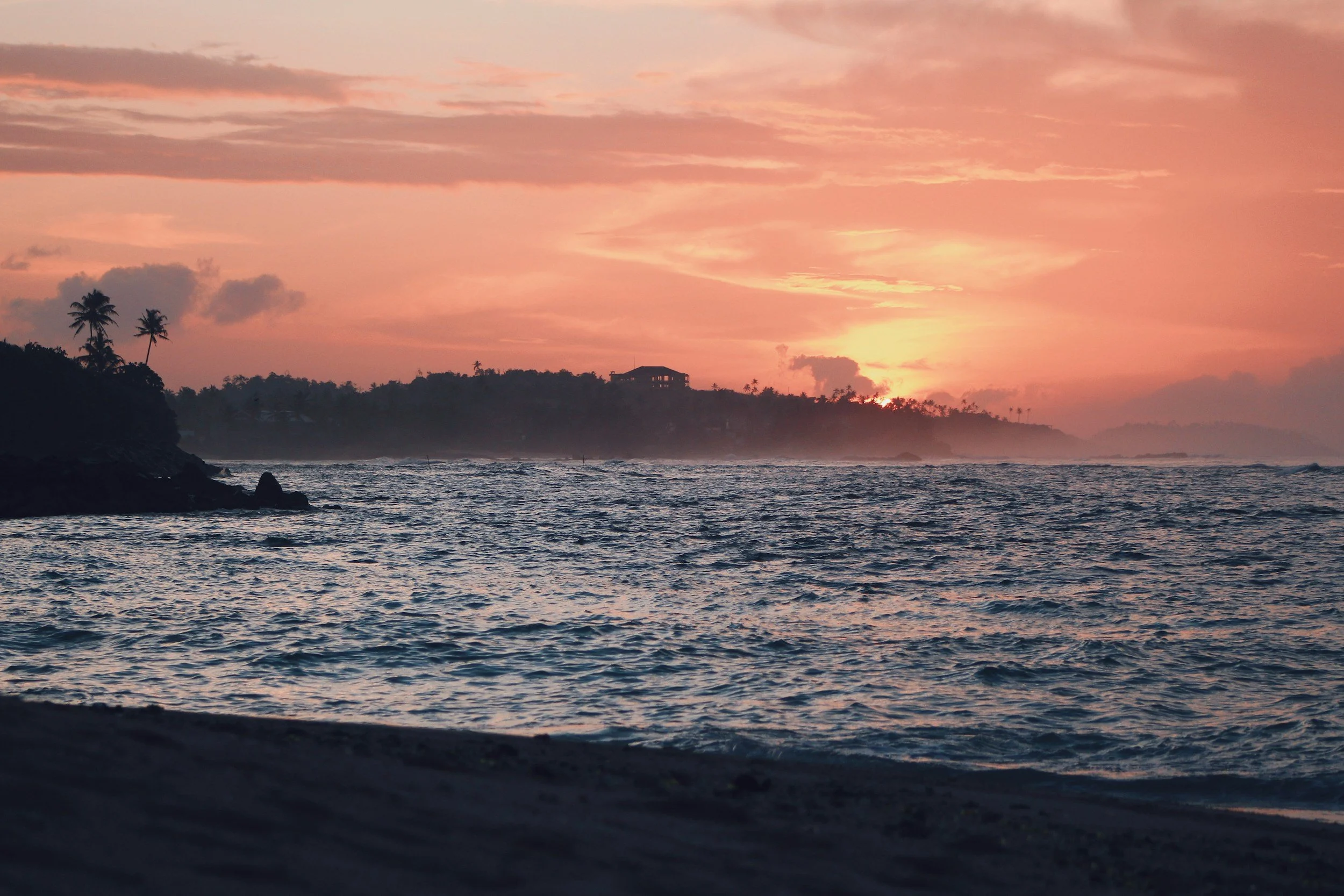 Sunset over the ocean with a pink and orange sky, silhouettes of palm trees on the shoreline, and a house on a distant hill.