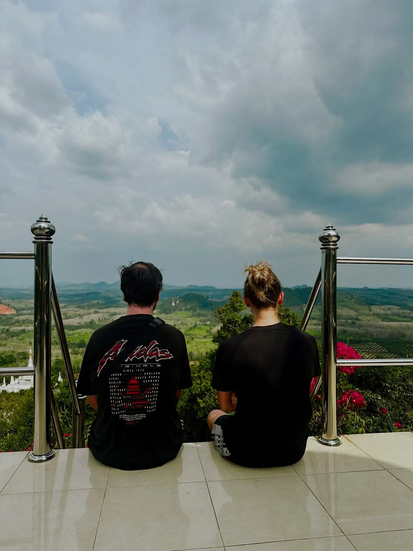 Two people sitting on a bench facing a scenic view of green hills and cloudy sky, with a metal railing and pink flowers nearby.