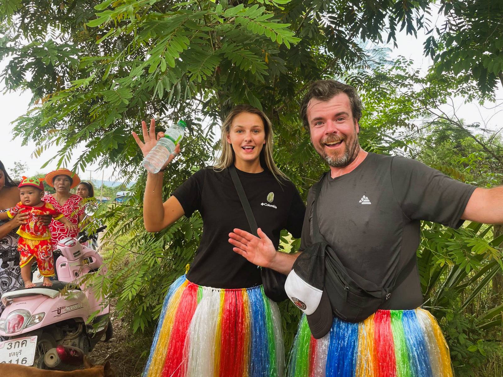 People taking a selfie outdoors in a lush, green environment. Two individuals are in the foreground wearing colorful grass skirts, with a woman holding a water bottle and a man smiling and waving. In the background, a small group of people, including a child dressed in a traditional outfit, and a pink scooter are visible.