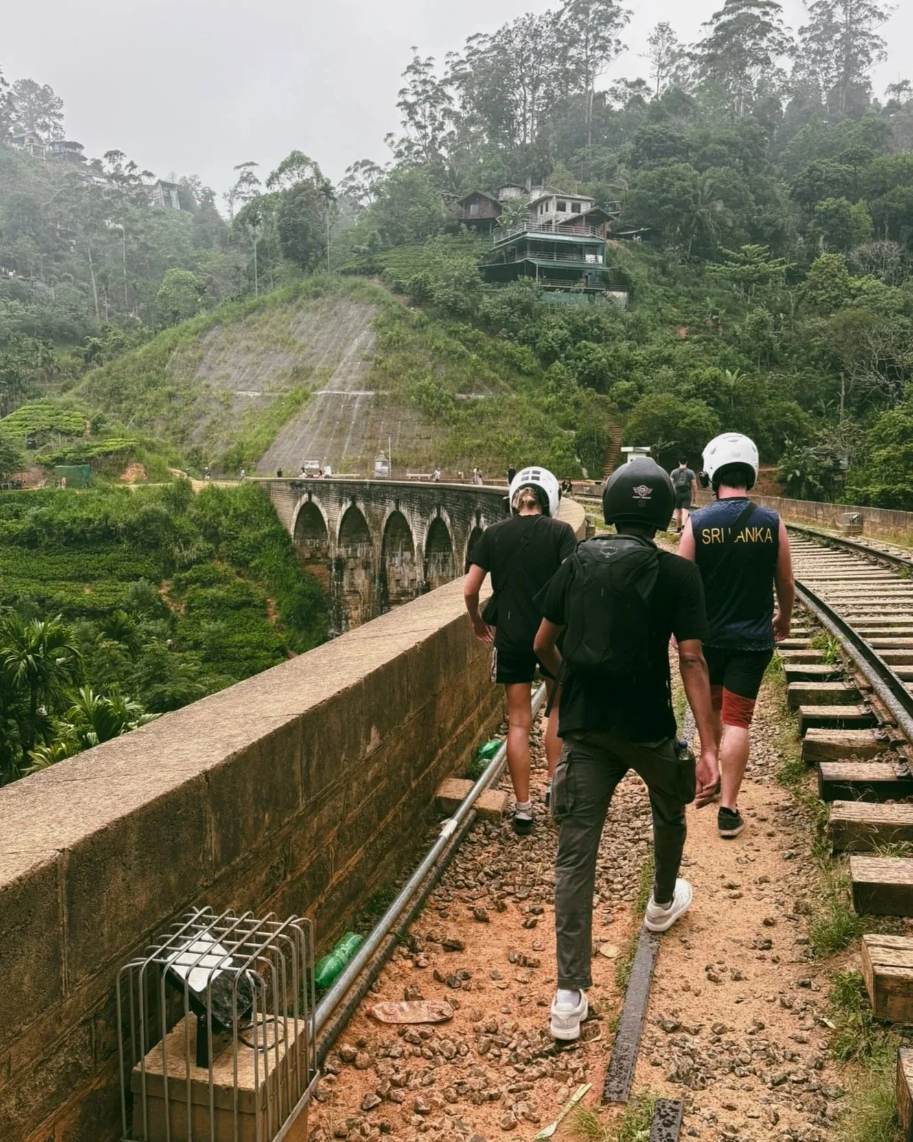 Three people walking along a railway track on a bridge in a lush green hilly area with houses on the hillside, overcast sky, some fog, wearing helmets and casual clothing.
