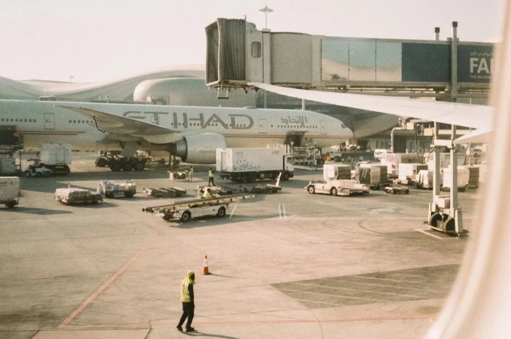Airplane at airport gate with ground crew working around it, jet bridge connected to aircraft, airport vehicles and luggage carts on tarmac.