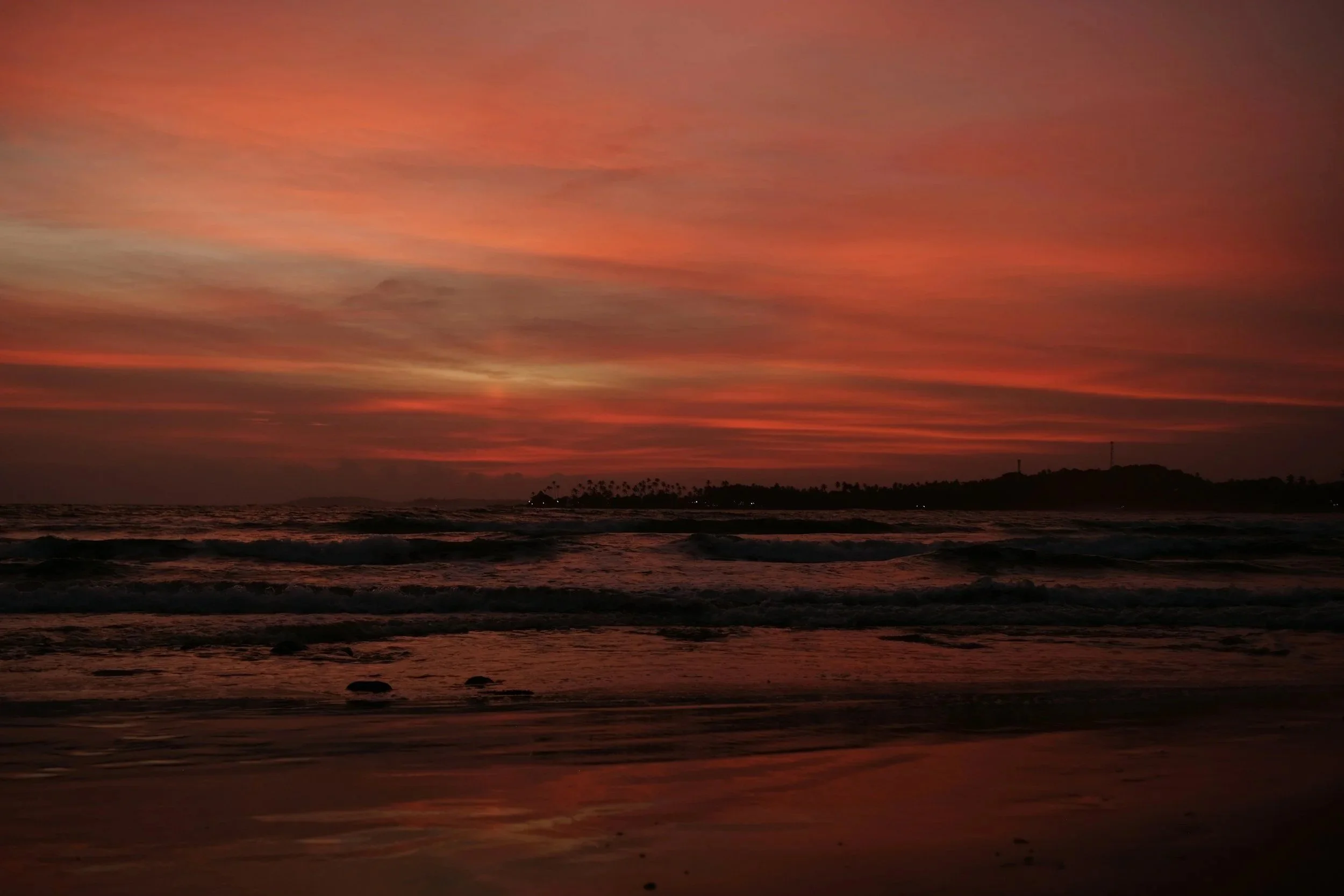 Sunset over the ocean with a colorful sky showing shades of pink, orange, and purple, visible clouds, and a silhouette of land in the distance.