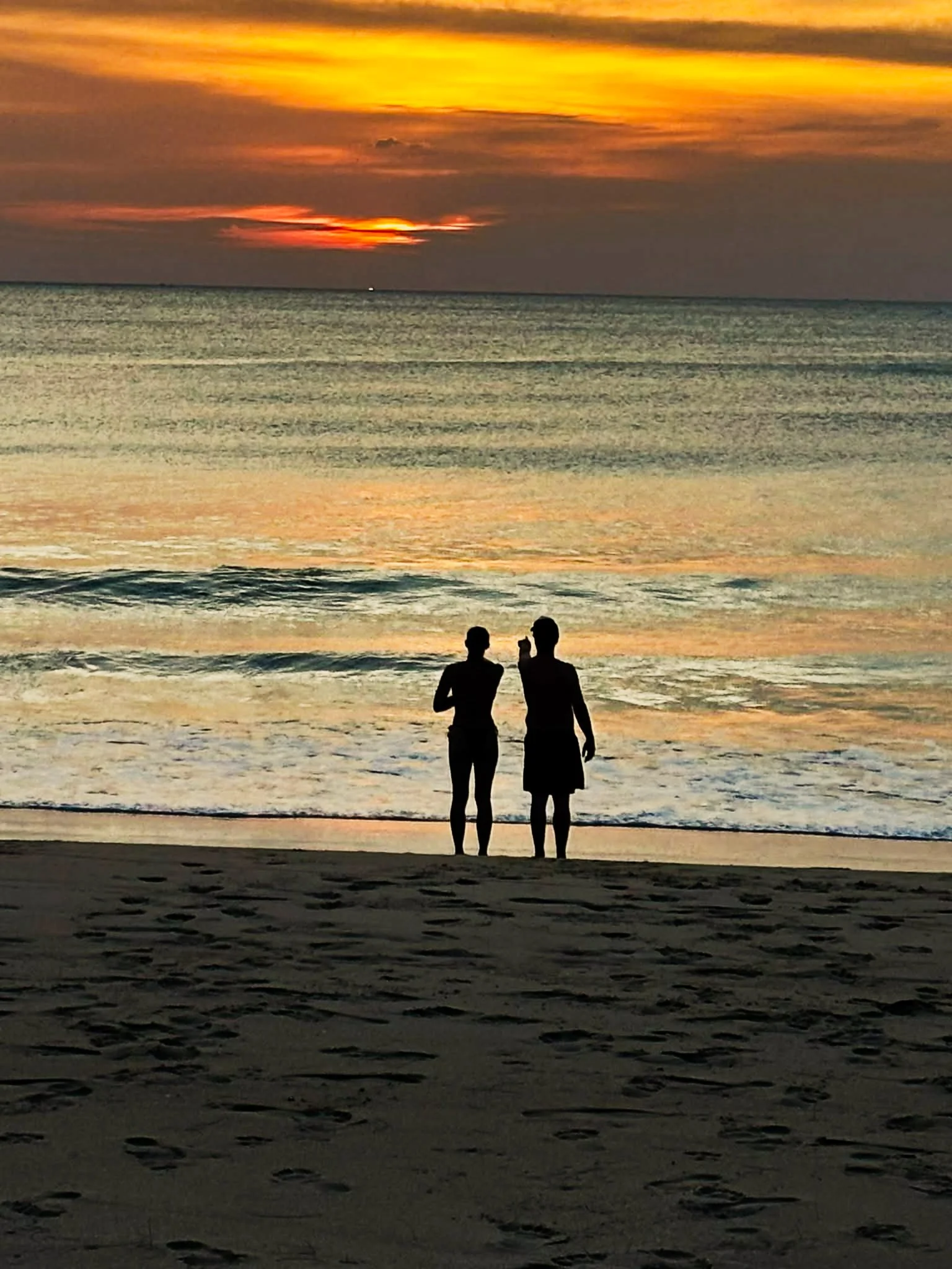 Two people standing on the beach at sunset, with the ocean and colorful sky in the background.