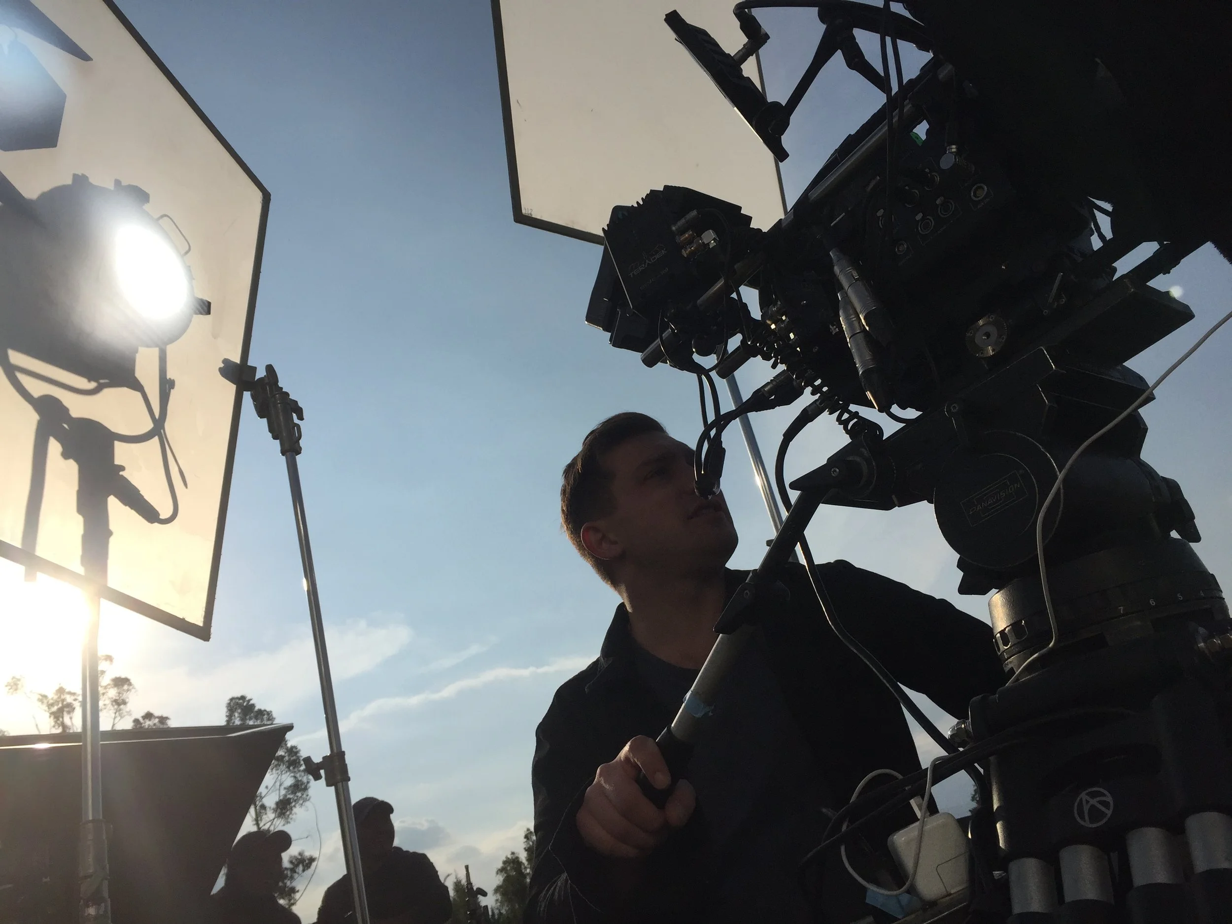 Behind-the-scenes shot of a camera operator filming outdoors, with professional lighting equipment and reflective screens visible against a clear blue sky.