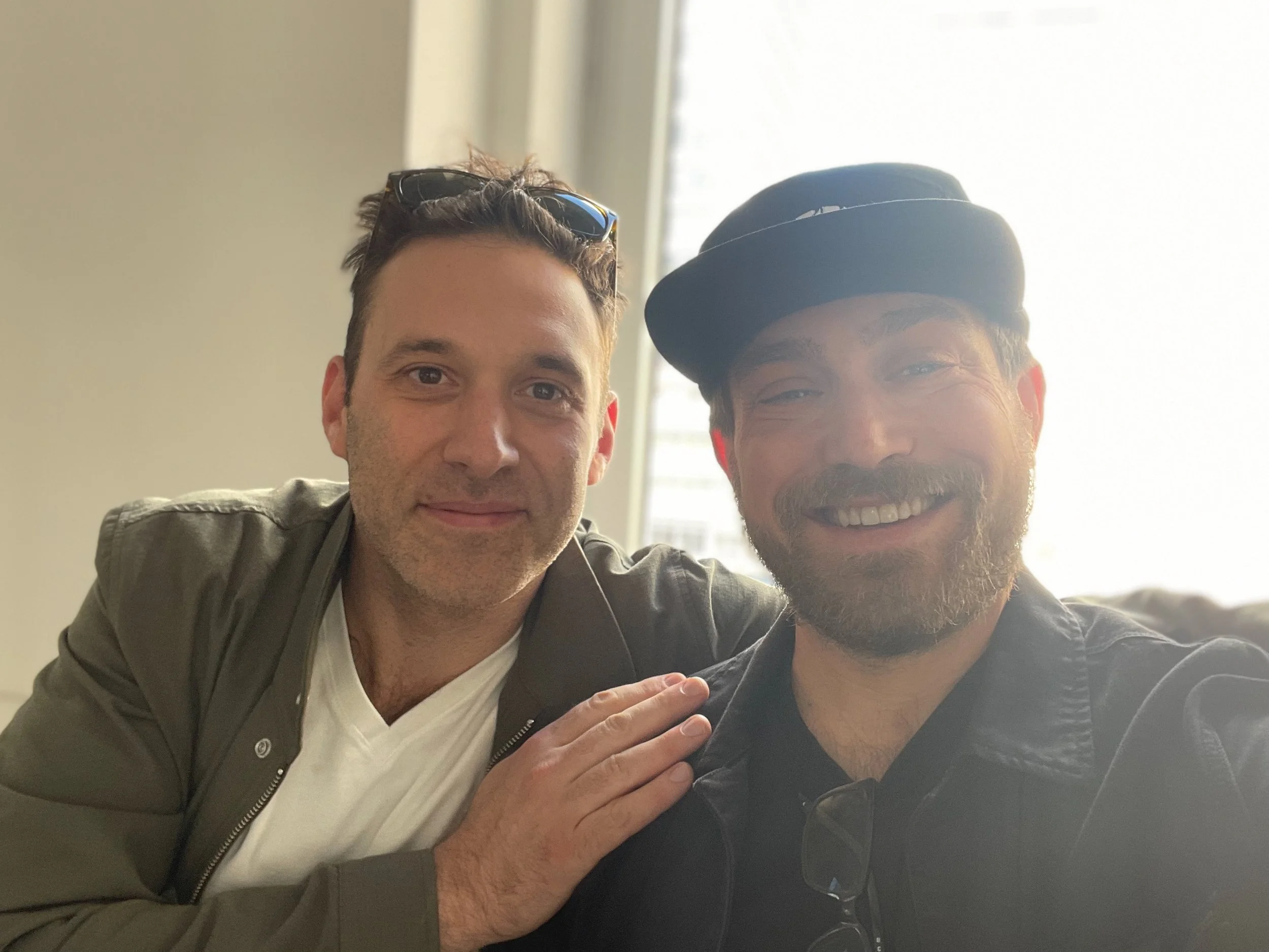 Two men posing for a photo indoors, with a window in the background. One man is wearing a jacket and sunglasses on his head, the other is wearing a cap and leather jacket. They are smiling and appear happy.