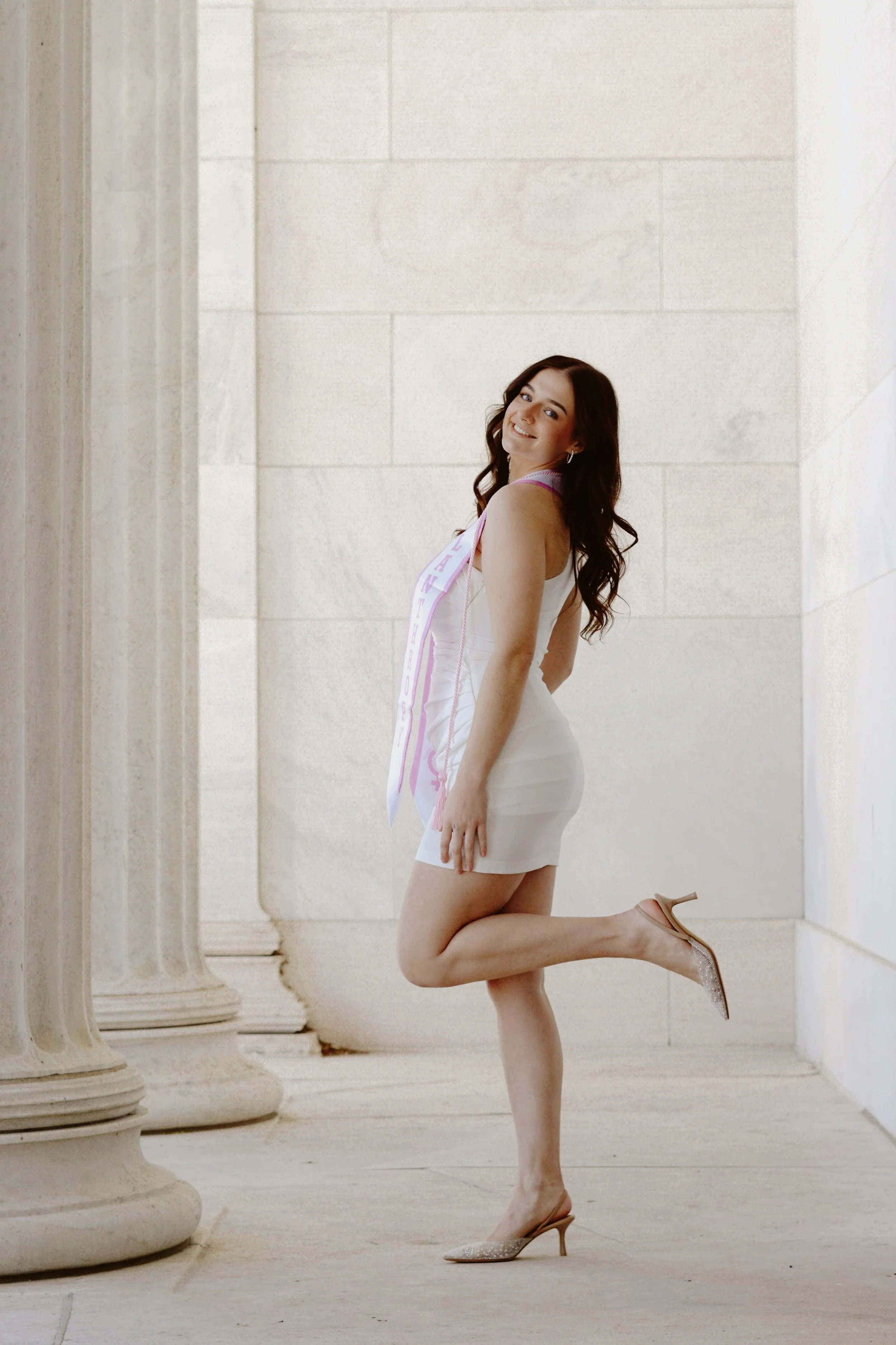 Young woman in white dress and high heels, wearing a sash, standing on one leg and smiling against a stone columned wall.