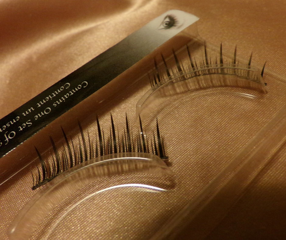 A pair of false eyelashes in clear plastic curved cases, with a box featuring an eye illustration partially visible in the background.