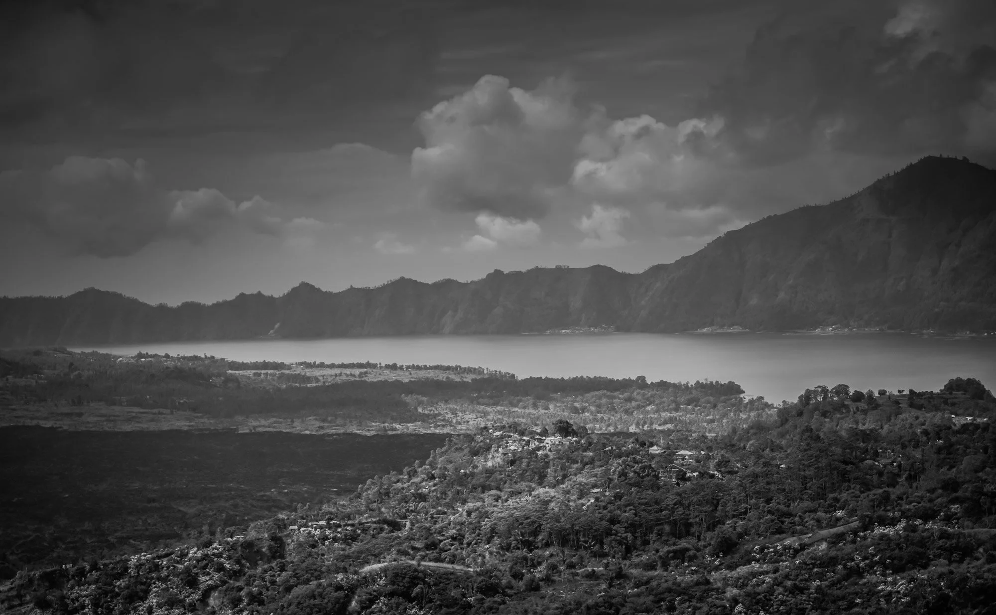 Black and white landscape of mountains, a lake, and forested hills under a cloudy sky.