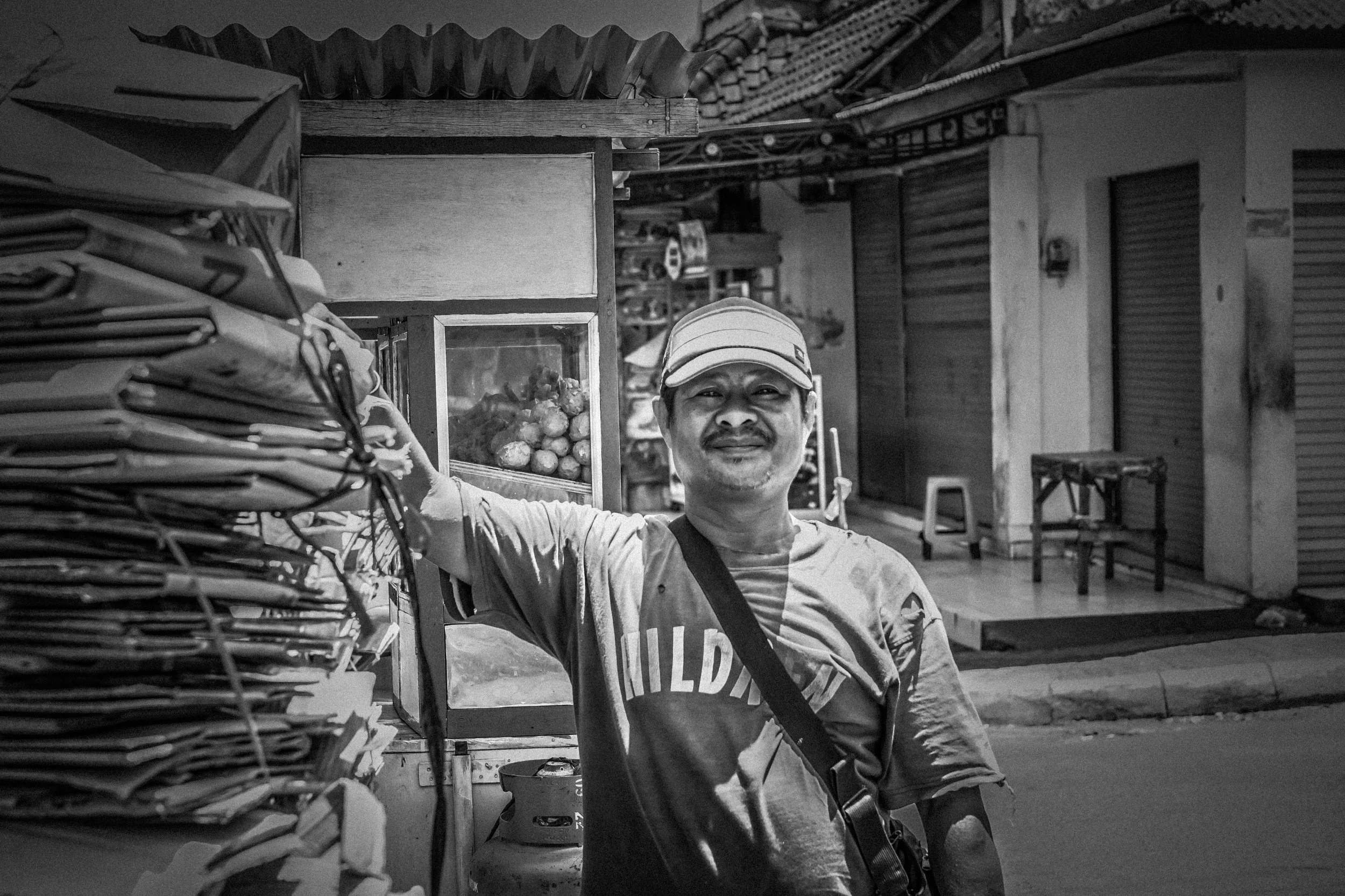 A man stands next to a small street cart selling newspapers and other items, smiling and wearing a cap, in an outdoor urban setting.