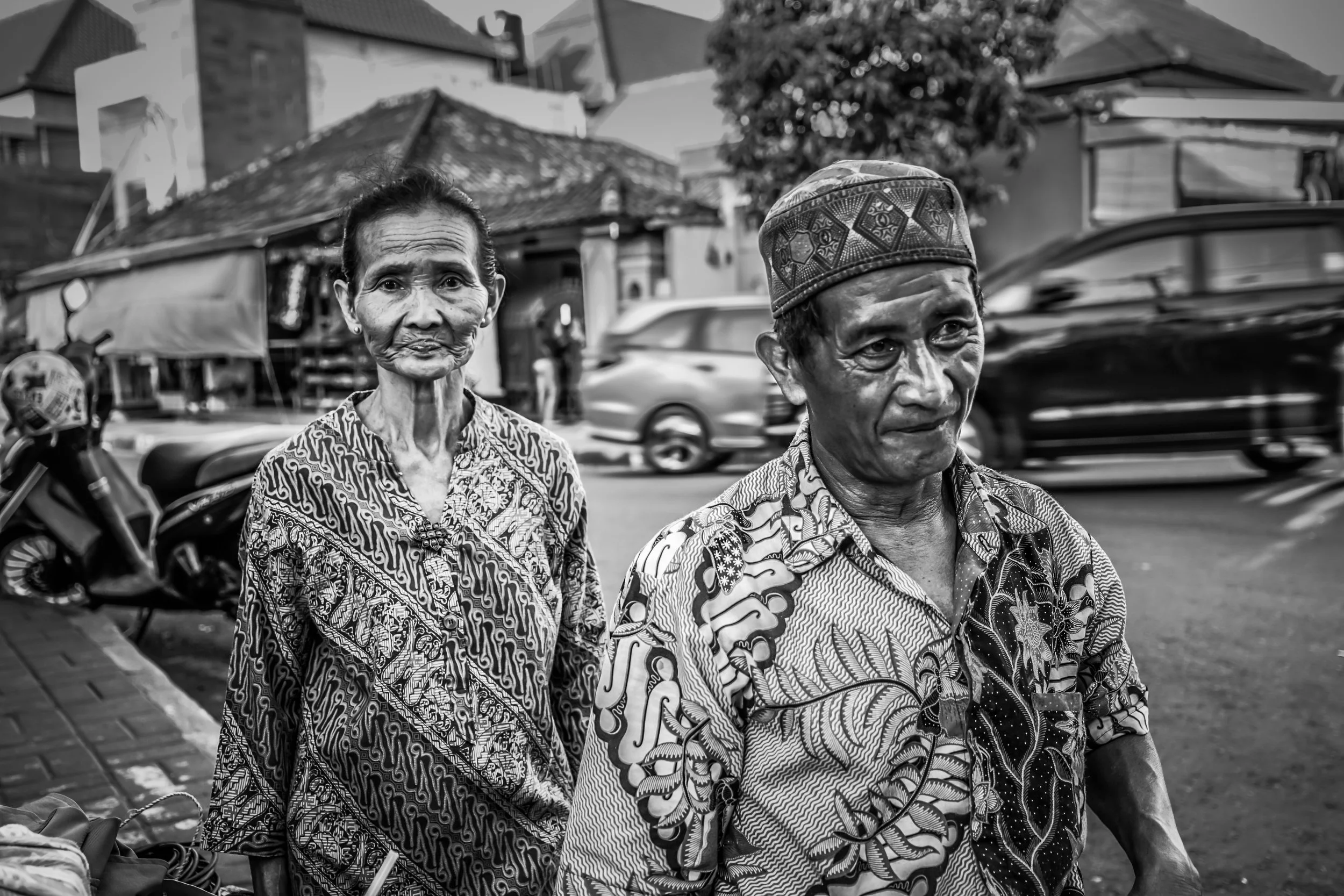 Black and white photo of an elderly man and woman standing on a busy street, with cars and a motorcycle passing by, in front of buildings with traditional architecture.
