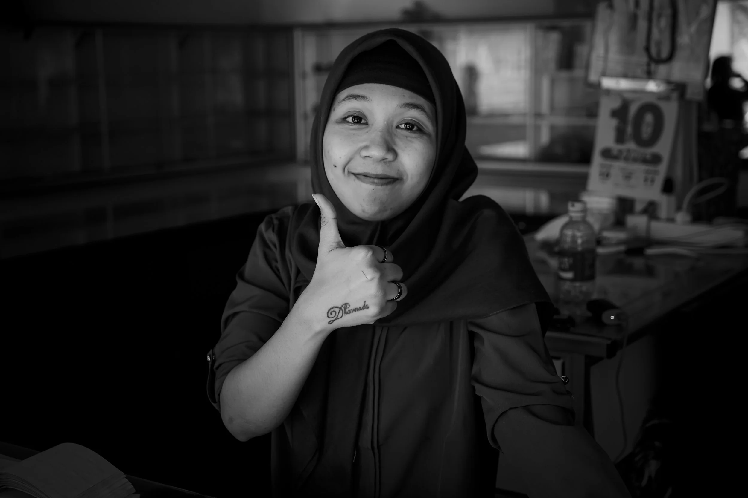 A young woman wearing a hijab giving a thumbs up and smiling at the camera in a black and white photo.