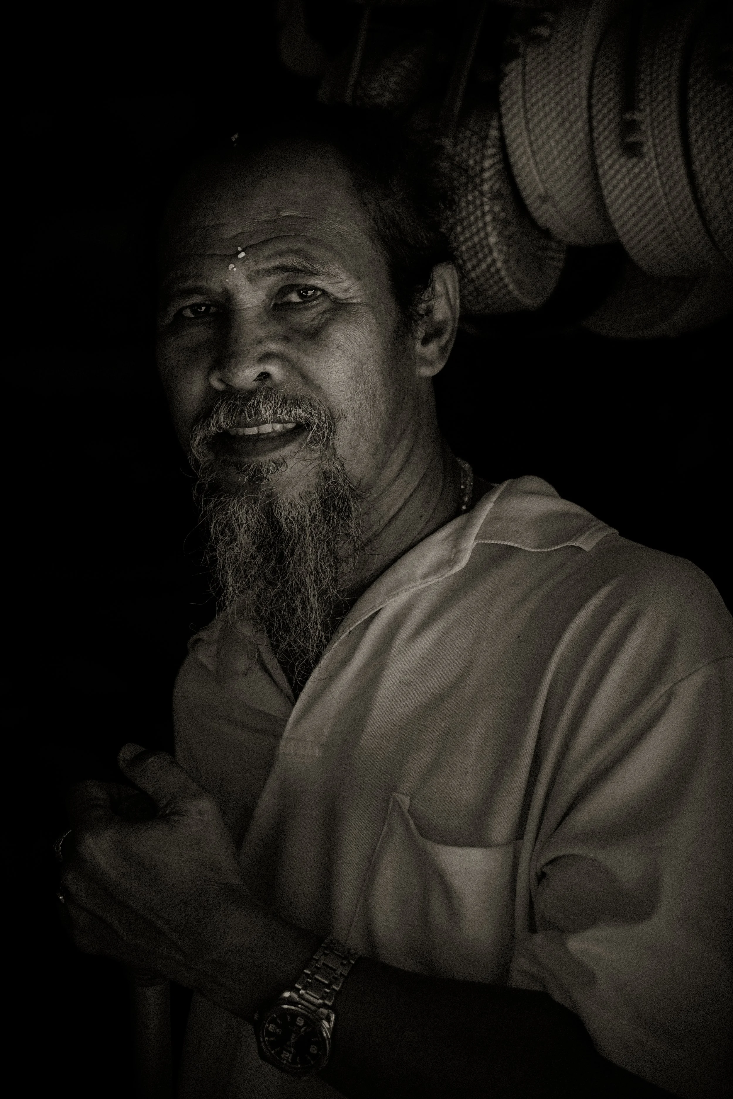 Black and white portrait of a middle-aged man with a beard and mustache, wearing a light-colored shirt, and a wristwatch, standing in a dimly lit space.