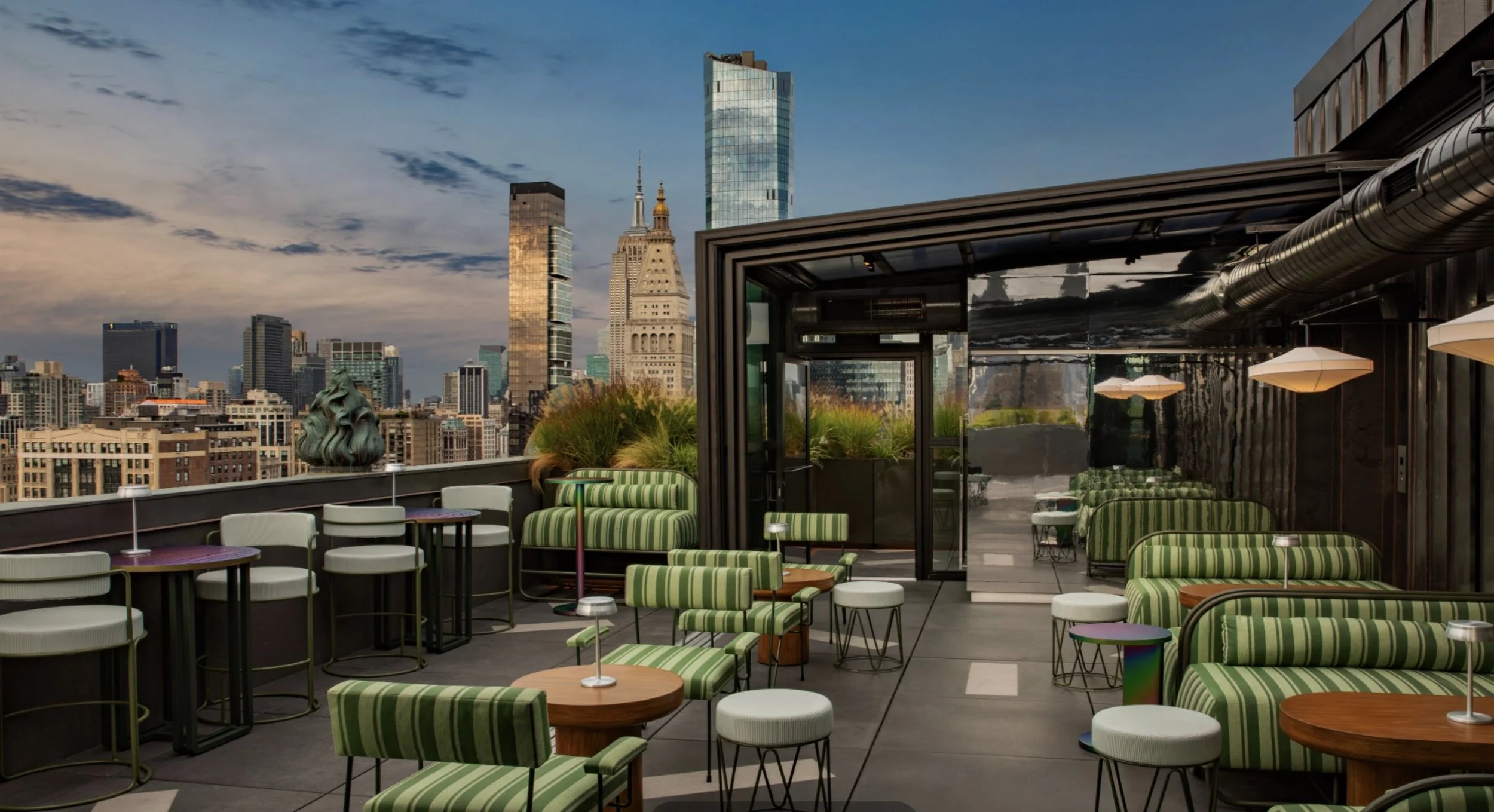 Rooftop bar with green striped chairs and tables, overlooking a city skyline with tall skyscrapers, during sunset.