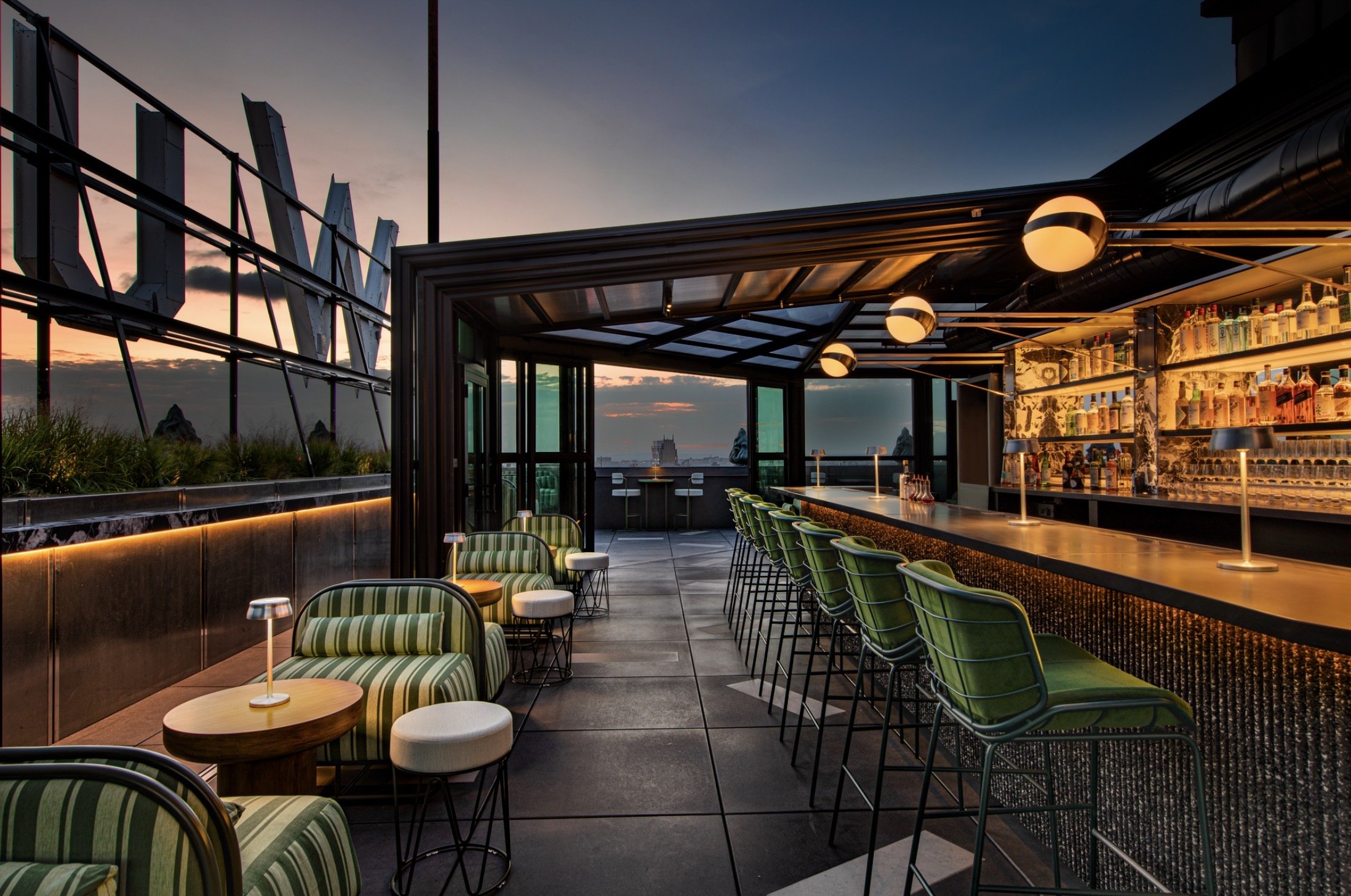 Rooftop bar with green striped chairs, a long bar counter, and a sunset view with city skyline in the background.