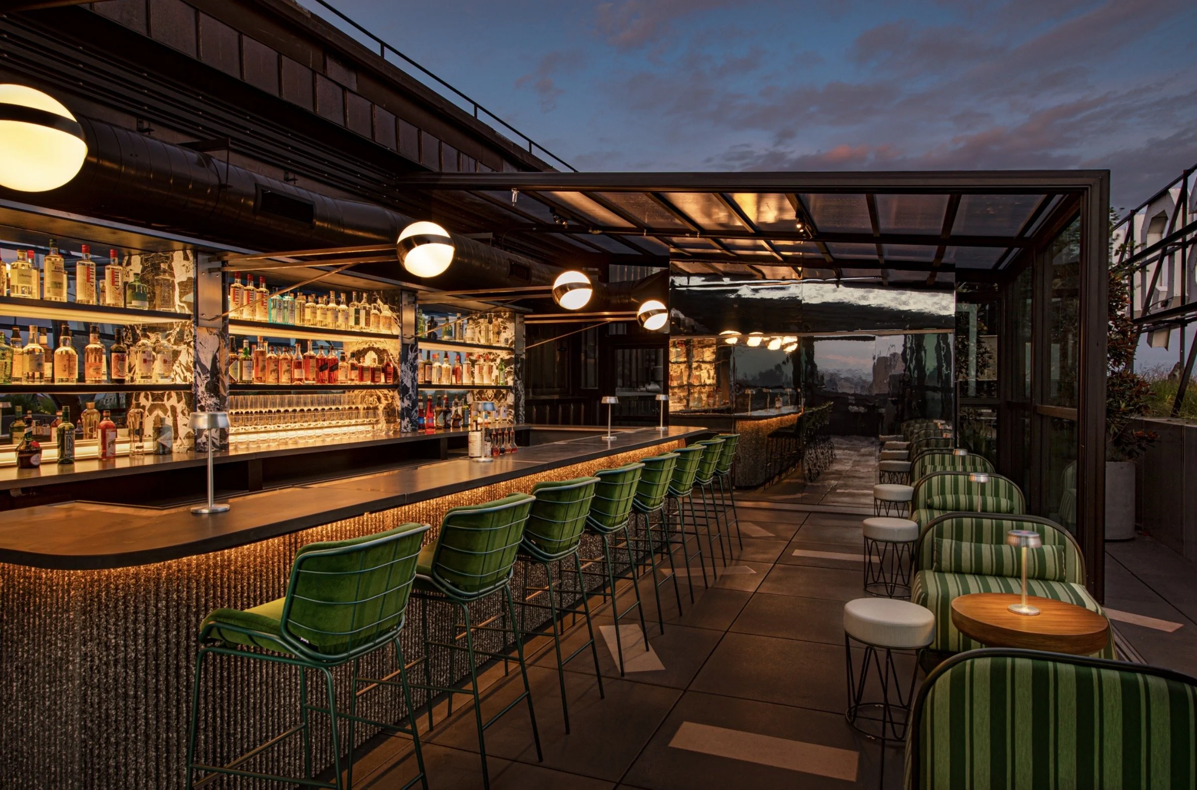Upscale rooftop bar with a fully stocked backlit liquor shelf and green chairs along the bar counter, featuring some lounge chairs with striped green upholstery and small tables with lamps, during sunset.