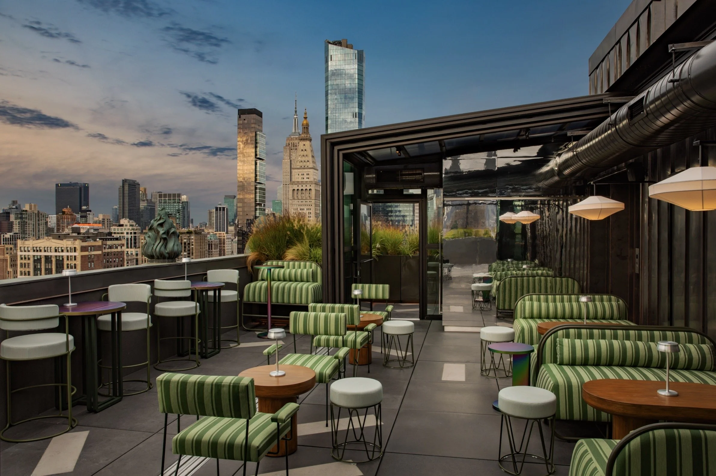 Rooftop patio with modern green and white striped chairs, small round tables with lamps, tall grasses in planters, and city skyline in the background under a partly cloudy sky.
