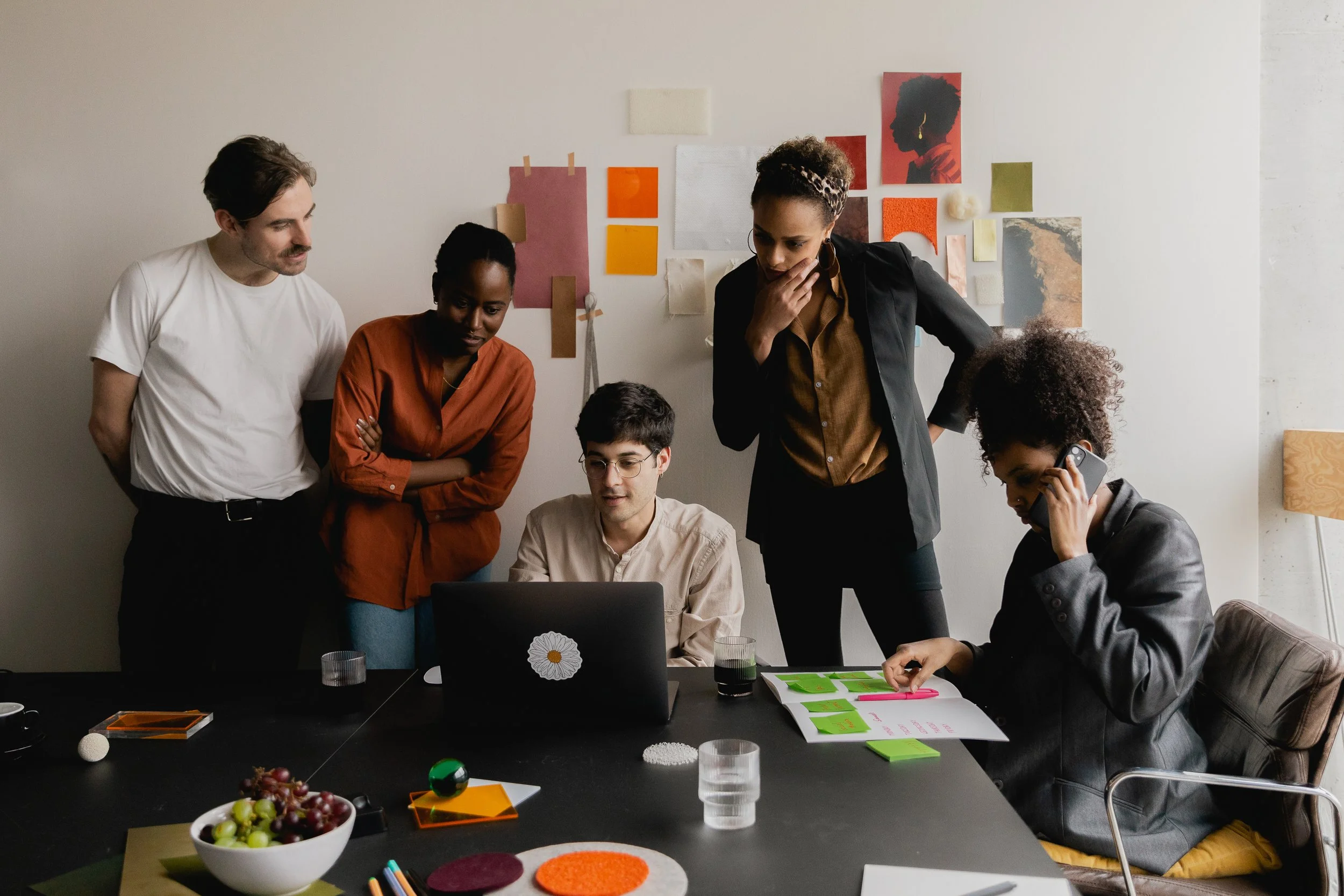 Five diverse people in a meeting room looking at a laptop and discussing, with various documents, a phone, and a bowl of fruit on the table.
