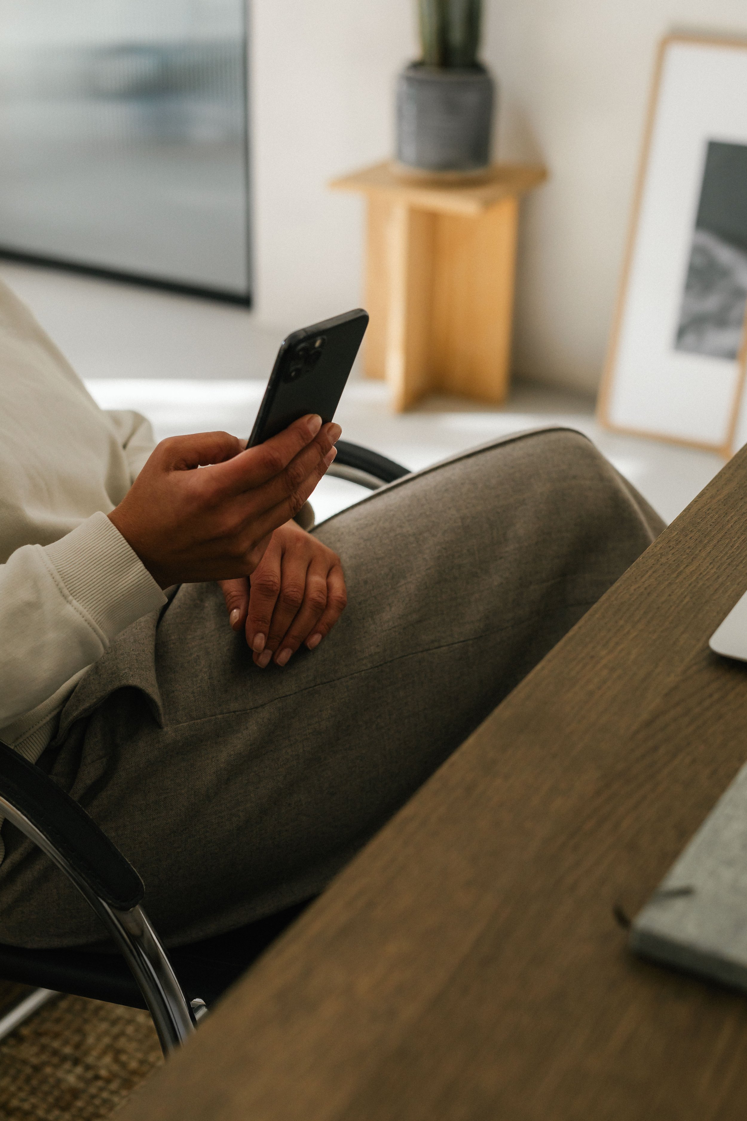 Person sitting on a chair at a desk, holding a smartphone in hand. A laptop is partially visible on the desk, with a framed photo or artwork and a potted plant on a small wooden side table in the background.