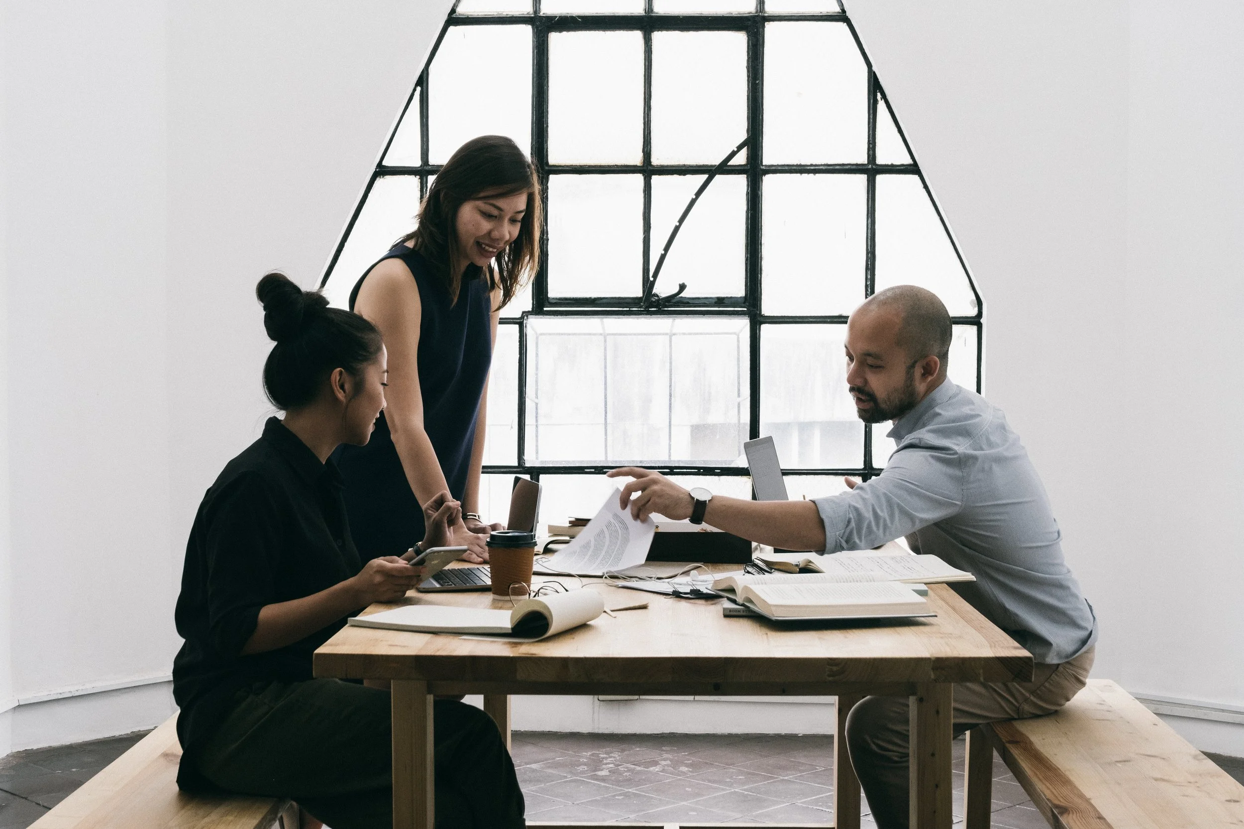 Three diverse people in an office having a discussion around a wooden table with laptops, notebooks, and a coffee cup.