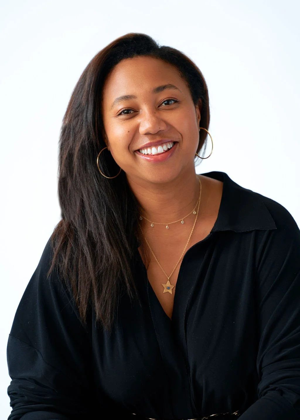 A smiling woman with long dark hair, wearing gold hoop earrings, a layered gold necklace with a star pendant, and a black top, against a plain white background.
