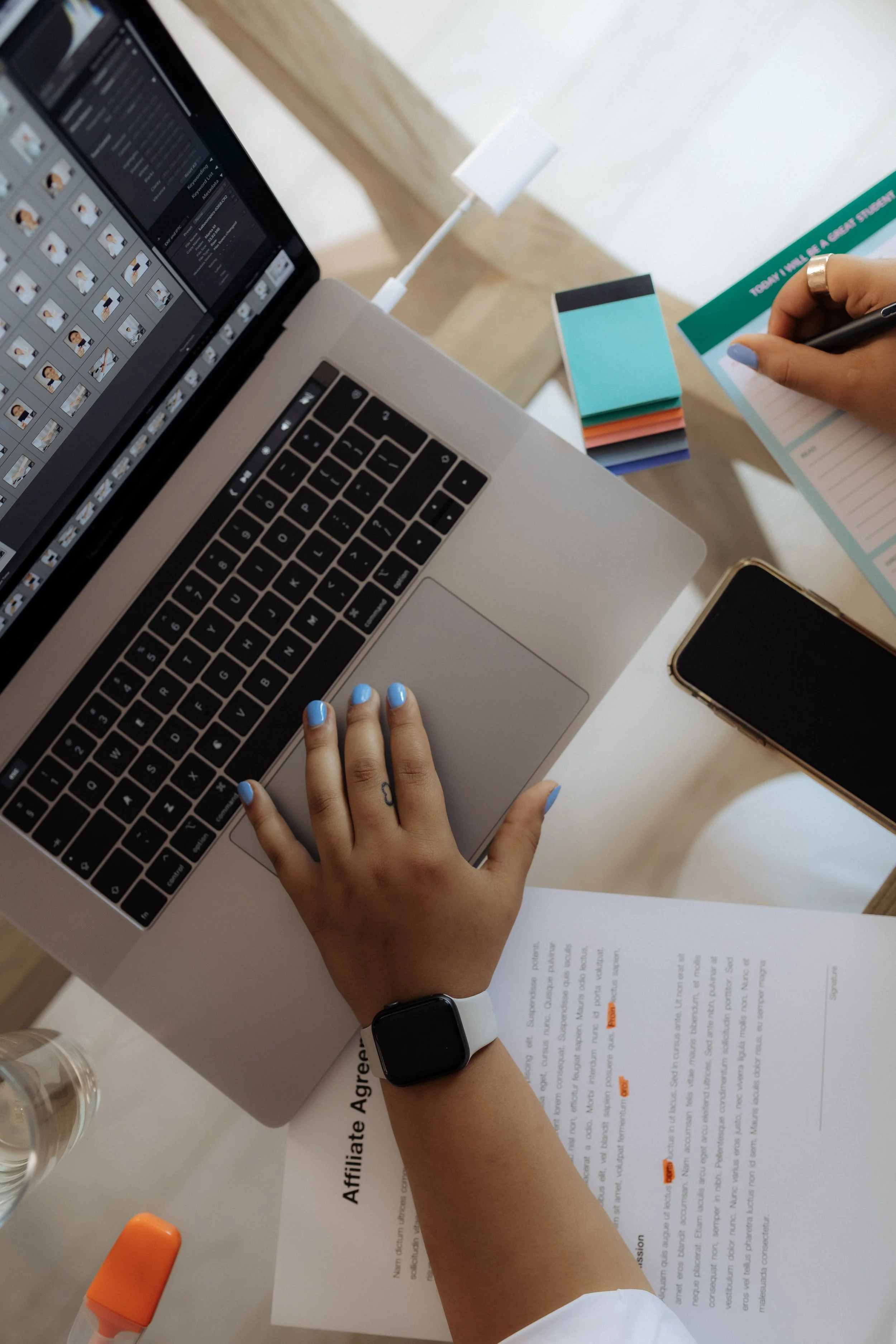 A person working on a laptop with a smartwatch, filling out an affiliate agreement form with stickers and a smartphone nearby.